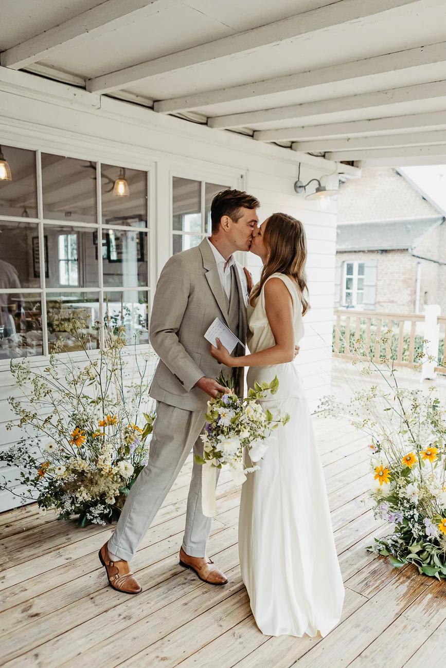 Jeunes mariés s'embrassant sur une terrasse en bois blanc, entourés de bouquets champêtres aux tons naturels.