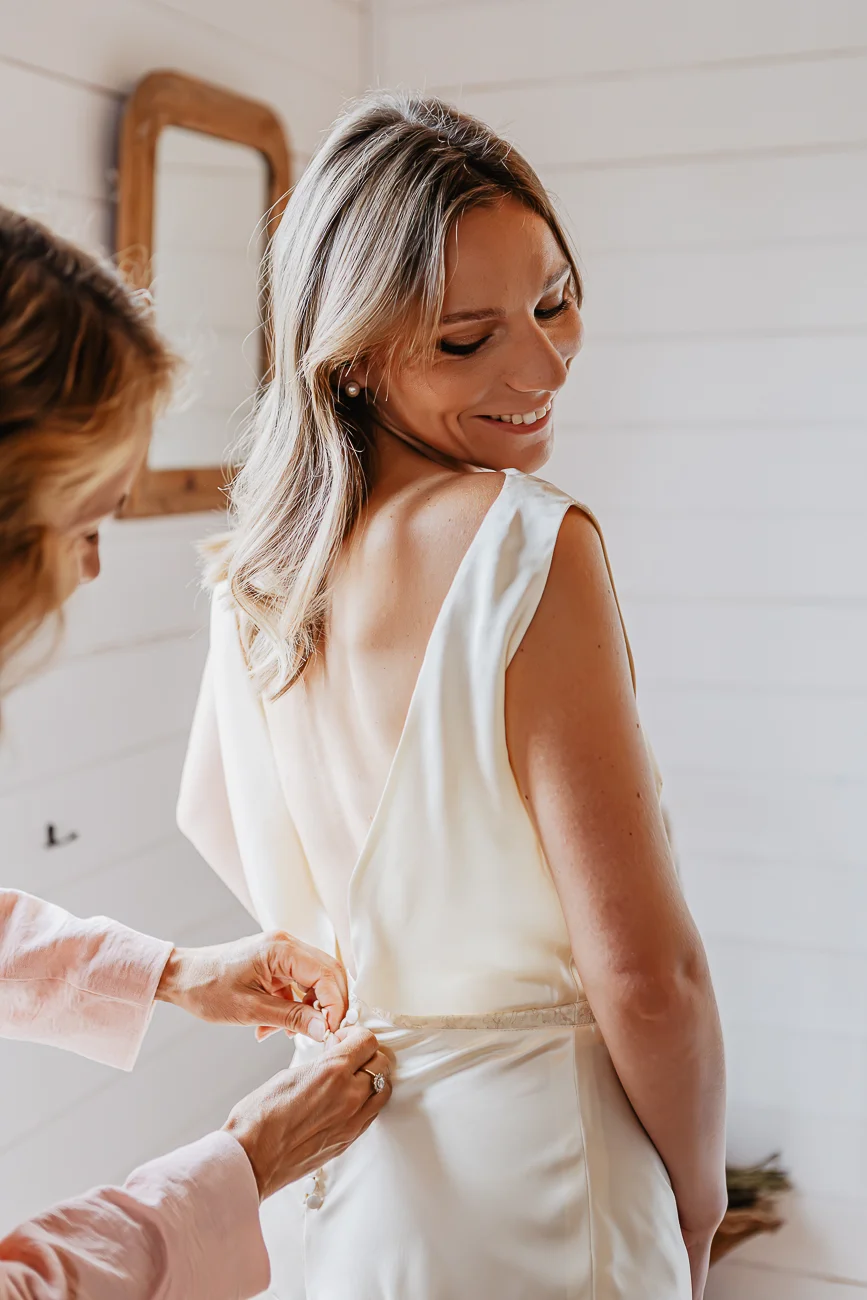 Une femme aide une mariée souriante à ajuster sa robe blanche satinée dans une pièce lumineuse et épurée.