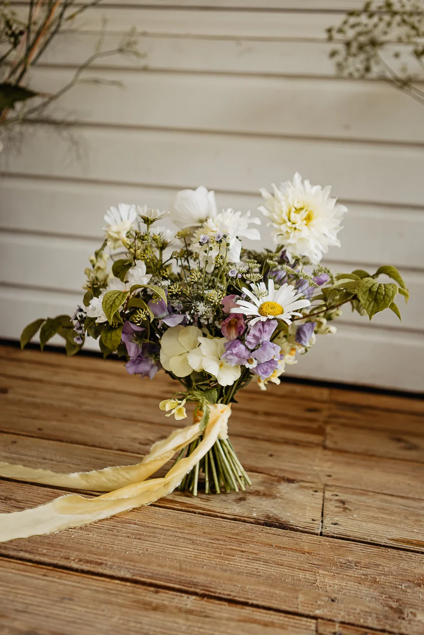 Bouquet de fleurs champêtres blanches et mauves, lié d'un ruban jaune, posé sur un parquet en bois rustique.