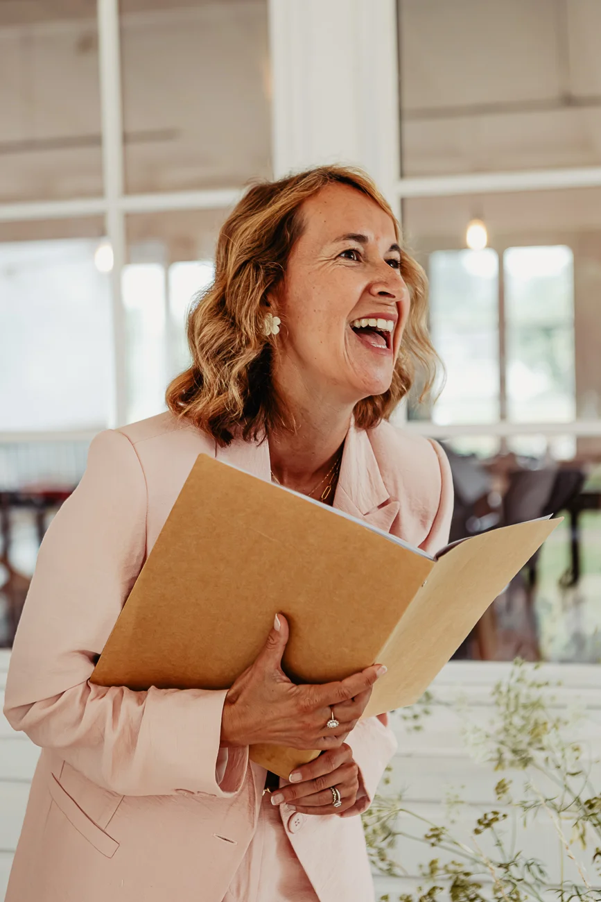 Une femme souriante en blazer rose pâle tenant un livret beige dans un intérieur lumineux.