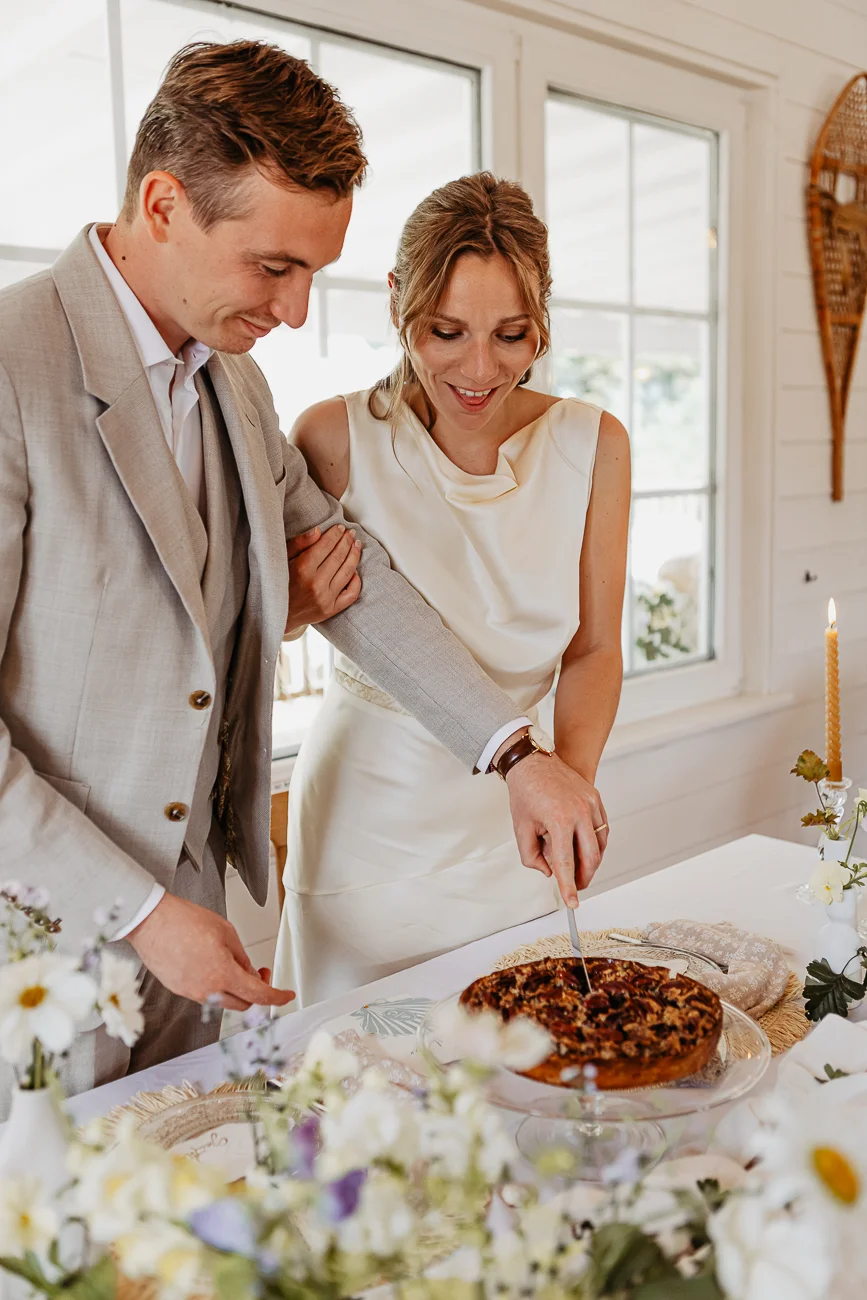 Un couple de mariés en tenues claires découpe ensemble un gâteau rustique lors d'une réception lumineuse et épurée.