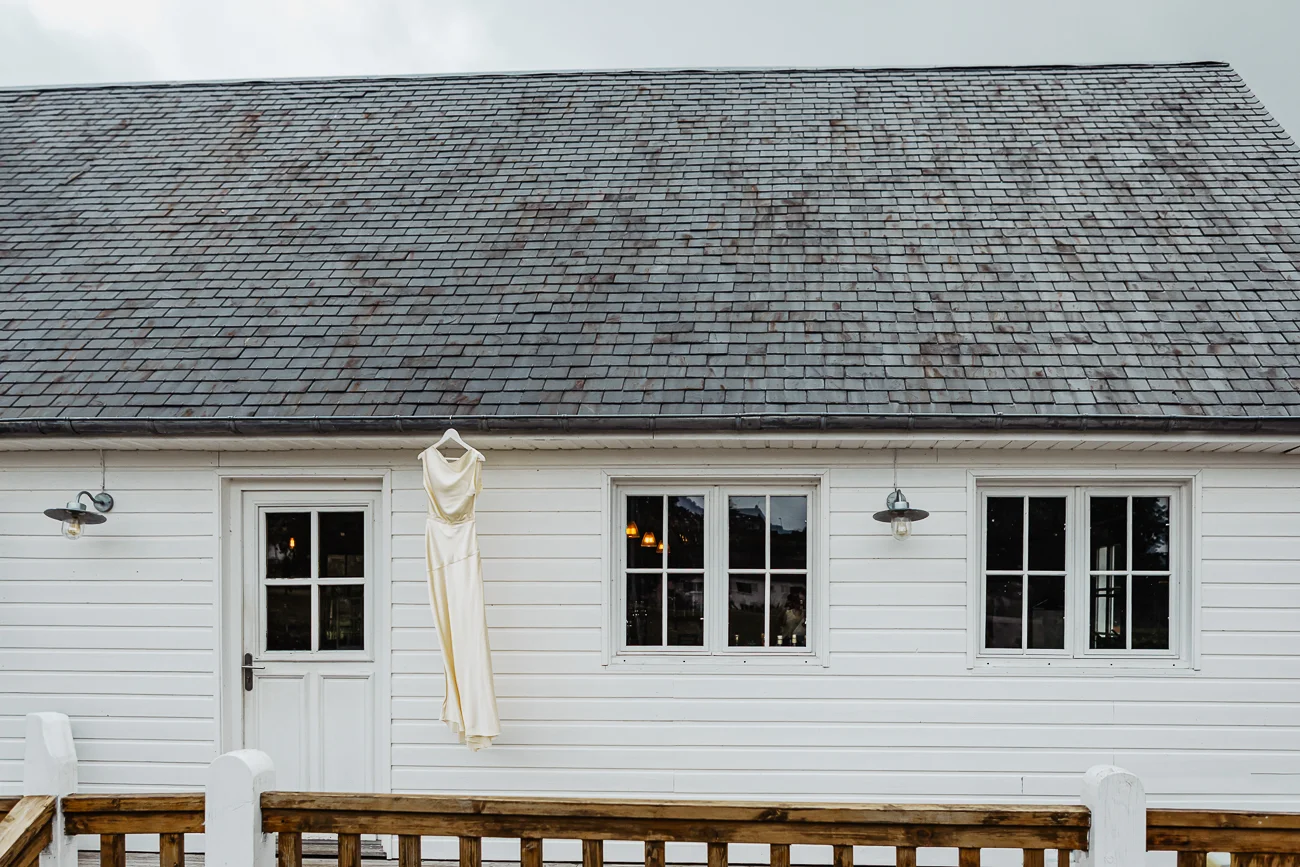 Robe de mariée blanche suspendue à la façade d'une maison en bois blanc sous un toit d'ardoises grises.