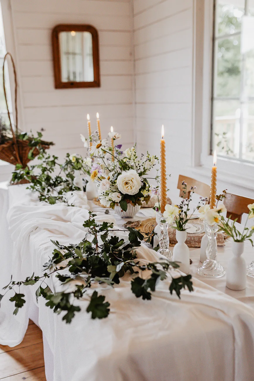 Table élégante dressée avec nappe blanche, compositions florales champêtres et bougies dans une ambiance lumineuse.