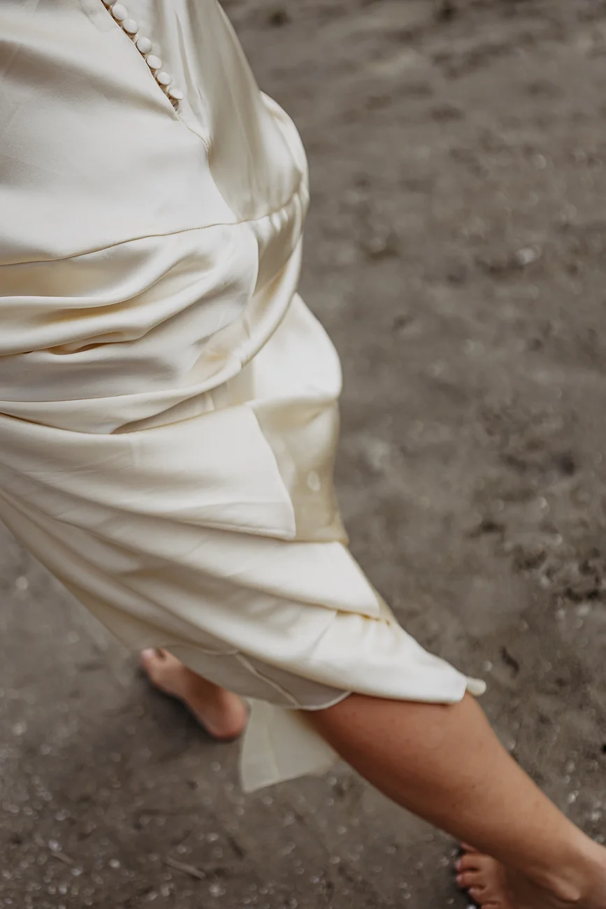 Gros plan sur une robe de mariée ivoire satinée et des pieds nus marchant sur le sable gris.