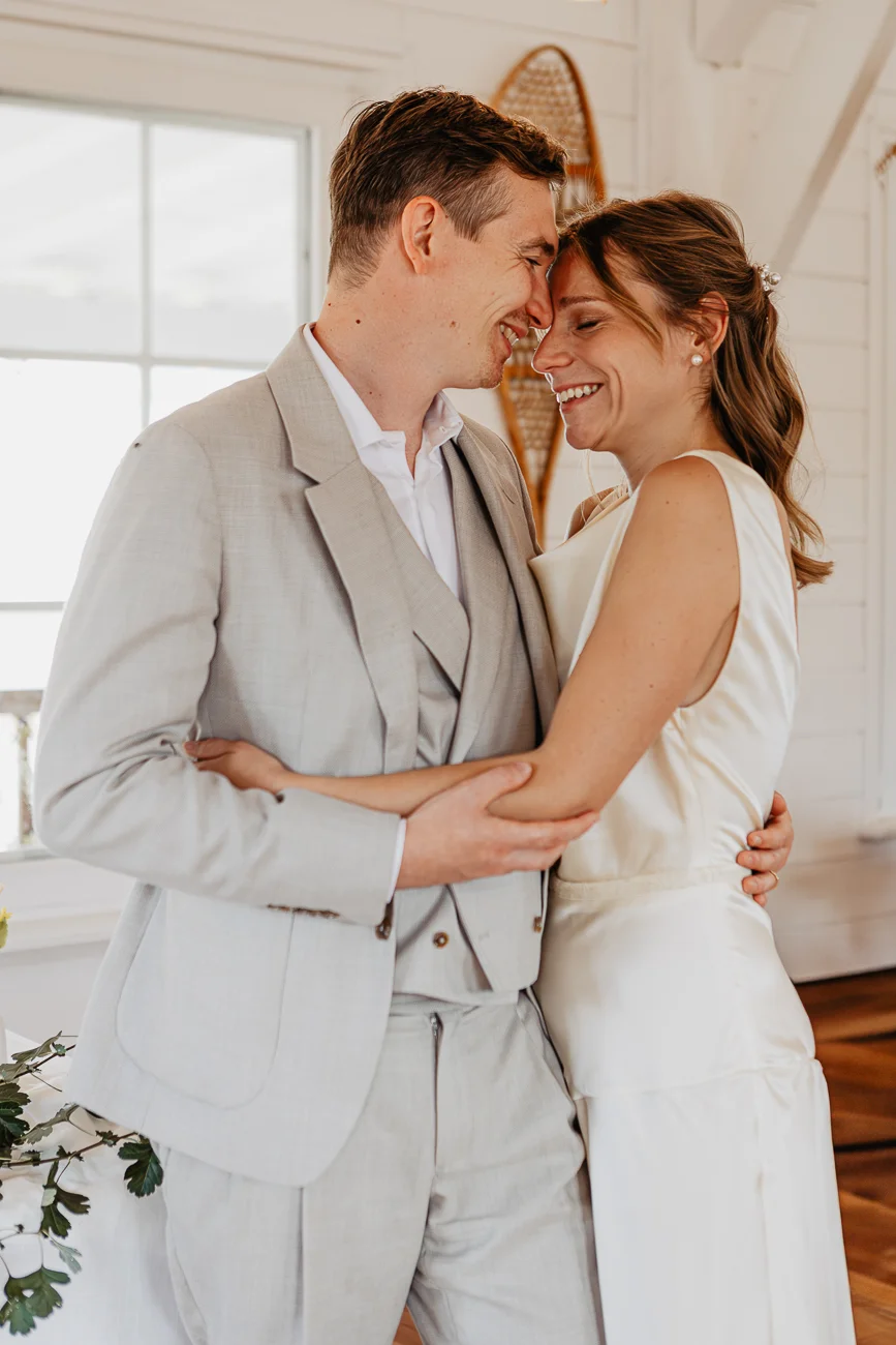 Couple marié complice et souriant, habillé de blanc et beige, s'enlaçant tendrement dans un intérieur lumineux.