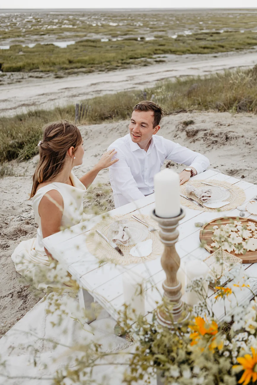 Couple en tenue blanche partageant un dîner romantique sur une plage de sable sous un ciel doux.