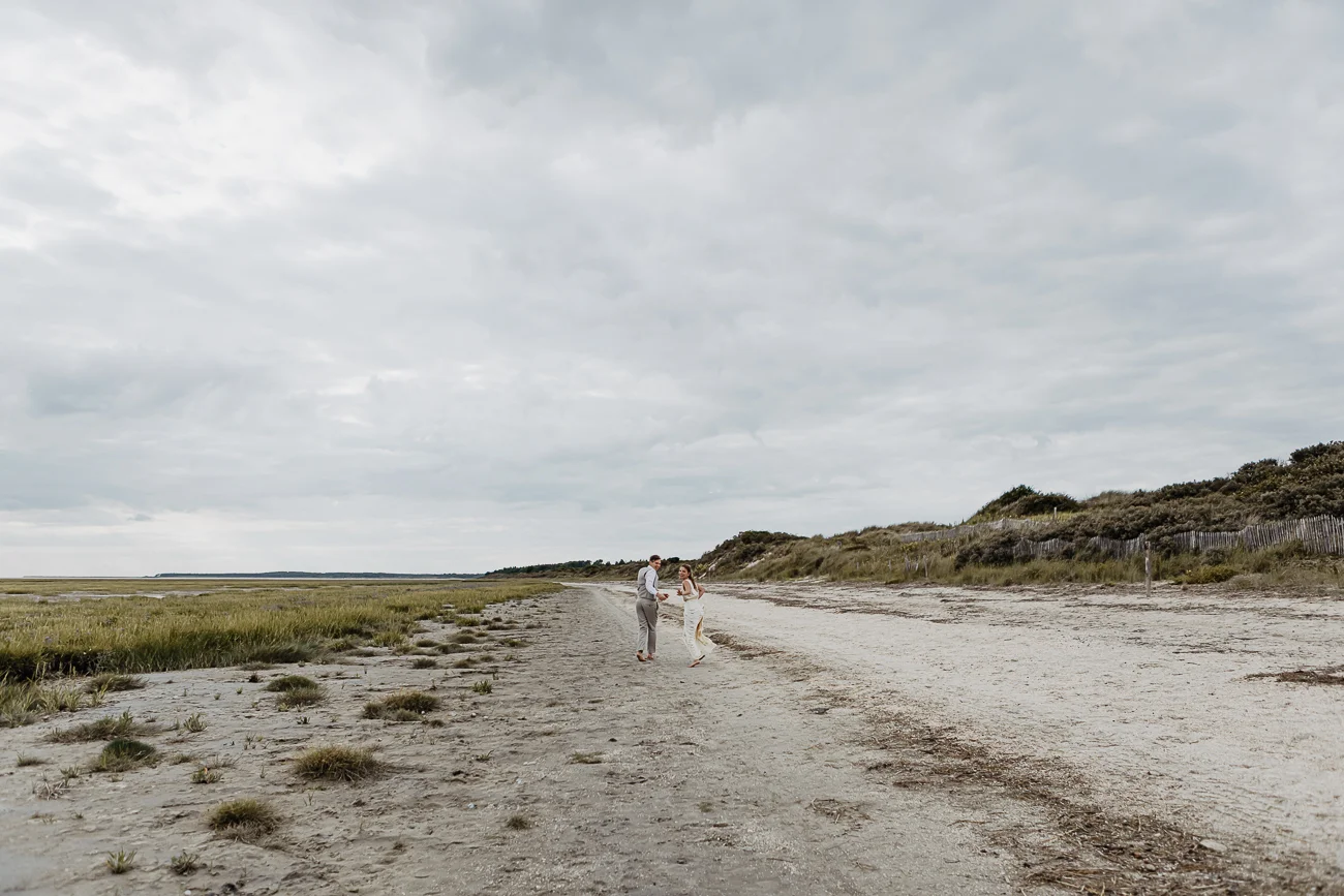 Un couple de mariés se tenant la main sur une plage sauvage et sablonneuse sous un ciel nuageux.