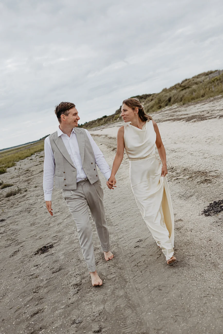 Jeunes mariés pieds nus marchant main dans la main sur une plage de sable gris sous un ciel nuageux.
