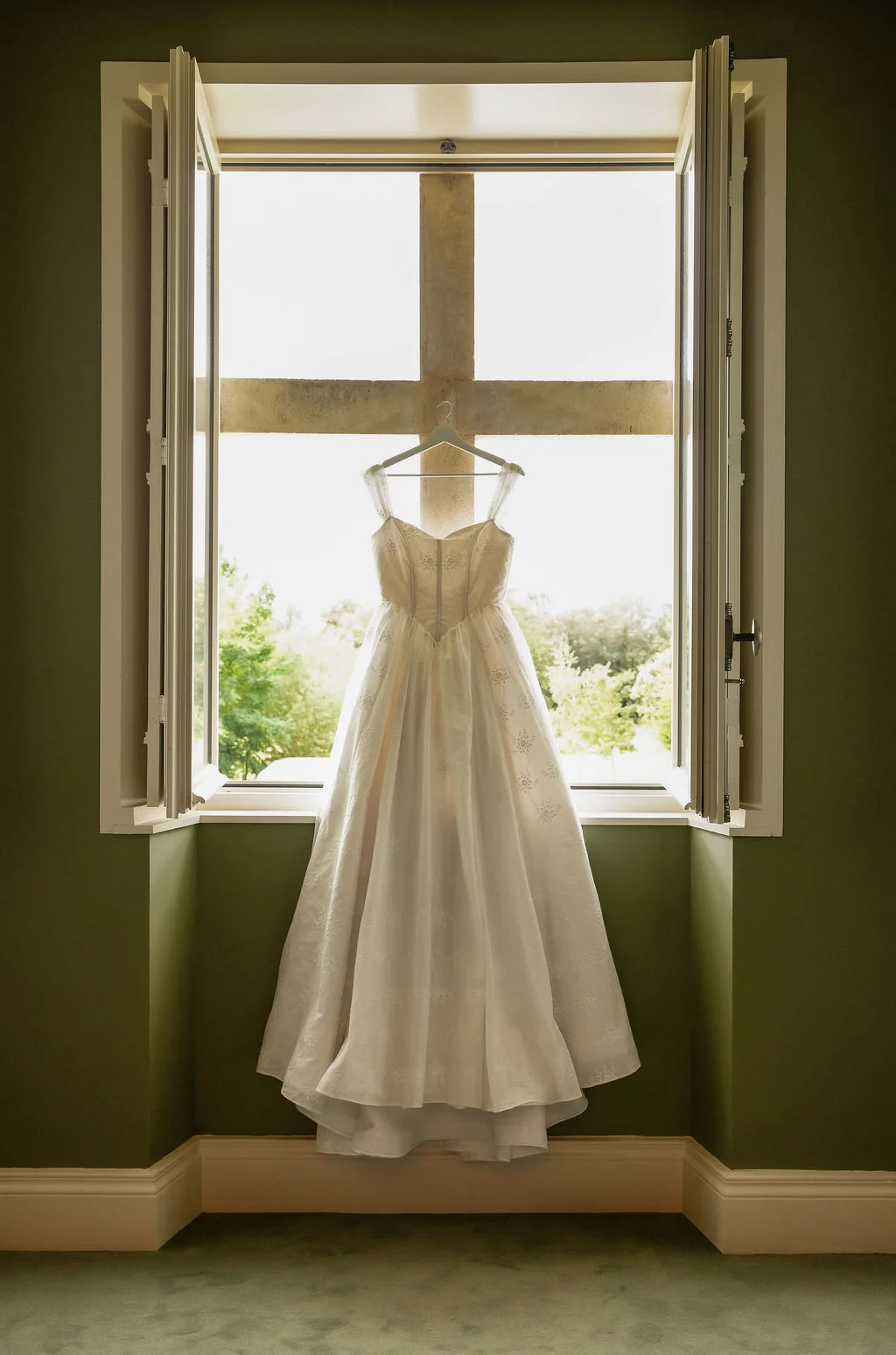 Robe de mariée blanche suspendue devant une fenêtre ouverte sur un jardin, dans une pièce aux murs vert olive.