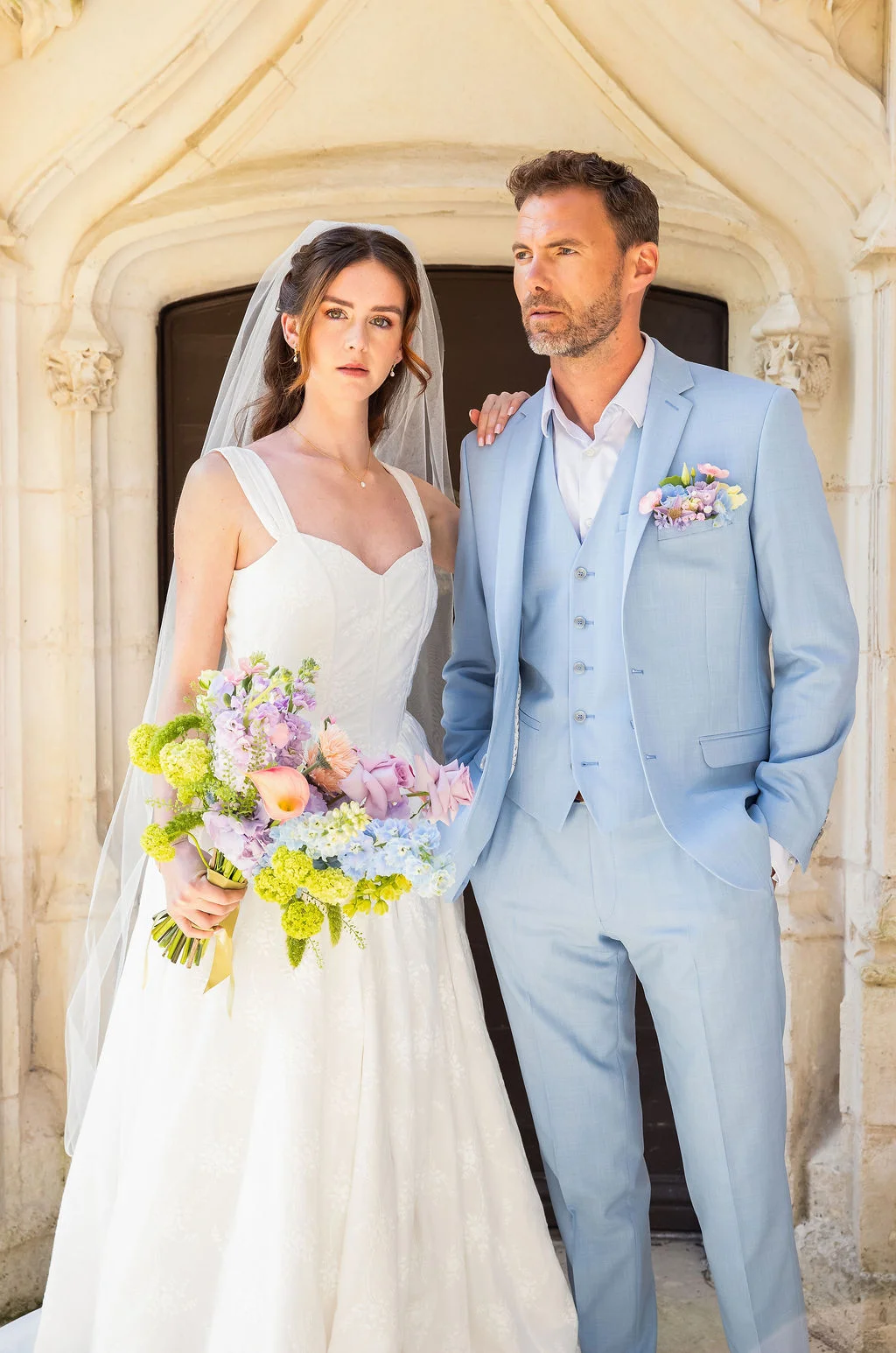 Couple de mariés élégants devant une porte ancienne, elle en robe blanche et lui en costume bleu ciel pastel.