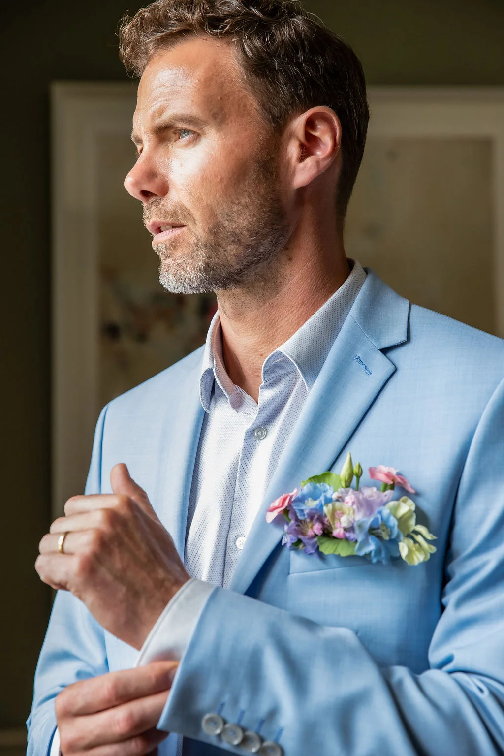 Homme élégant en costume bleu ciel avec une boutonnière fleurie, regardant au loin dans une ambiance romantique.