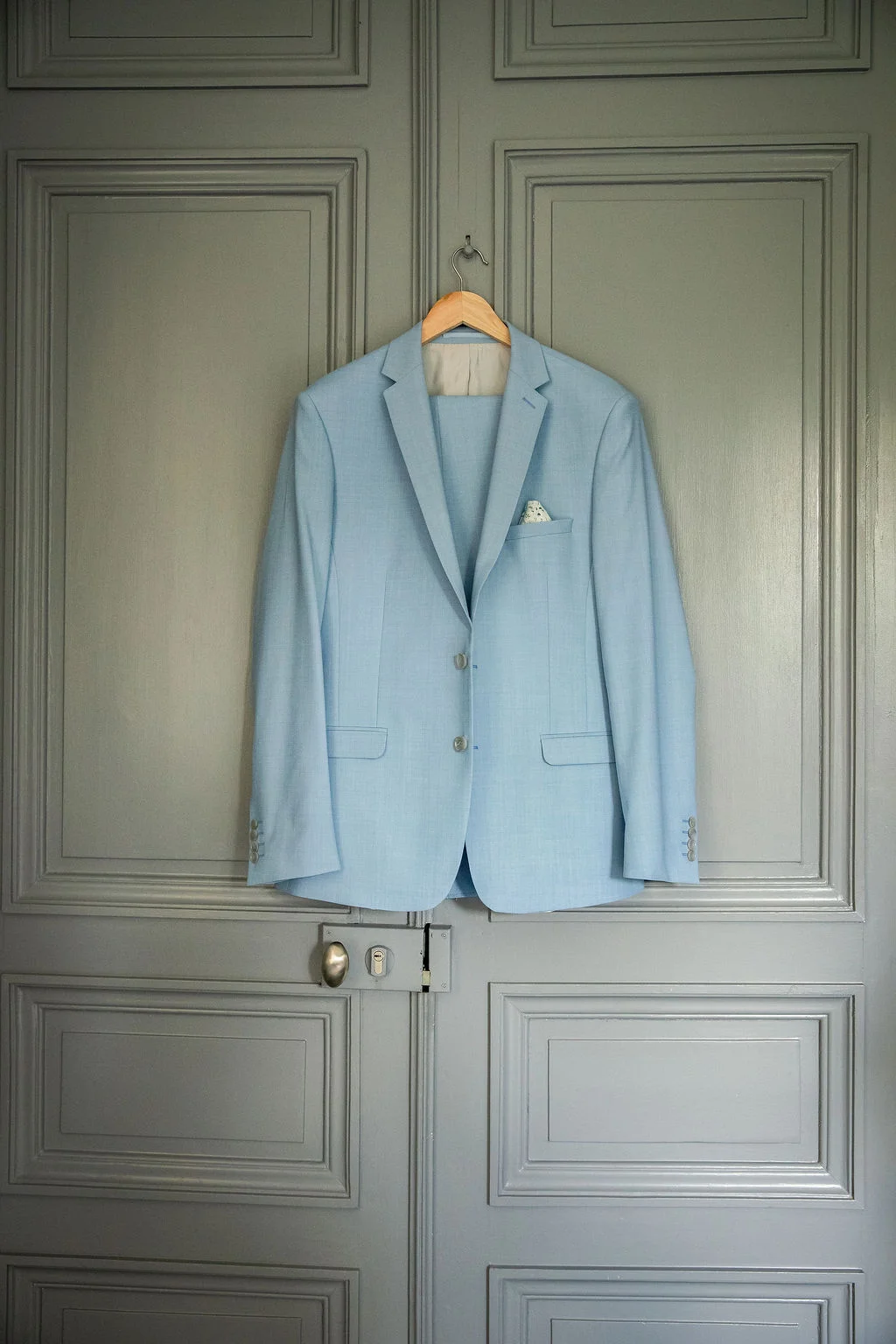 Costume de mariage bleu ciel suspendu à une porte grise, ambiance romantique et lumineuse.