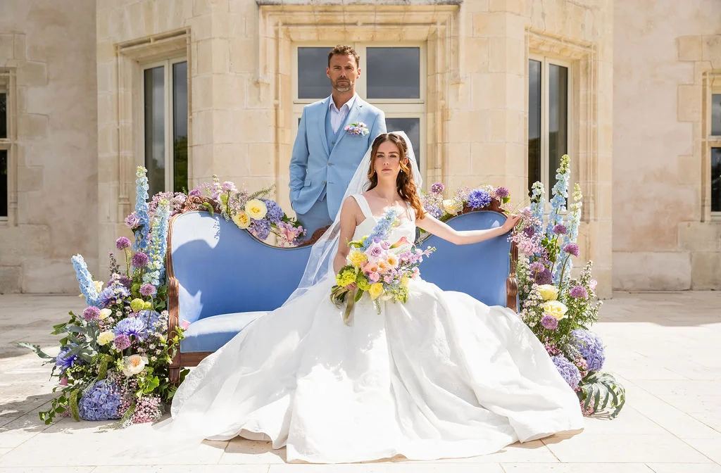 Couple de mariés élégants posant sur un canapé bleu entouré de fleurs colorées devant un château en pierre claire.