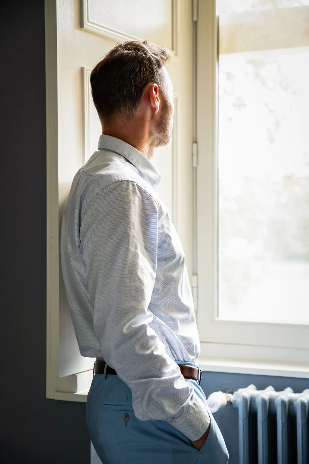 Homme de profil en chemise claire et pantalon bleu, regardant par la fenêtre dans une ambiance lumineuse et sereine.