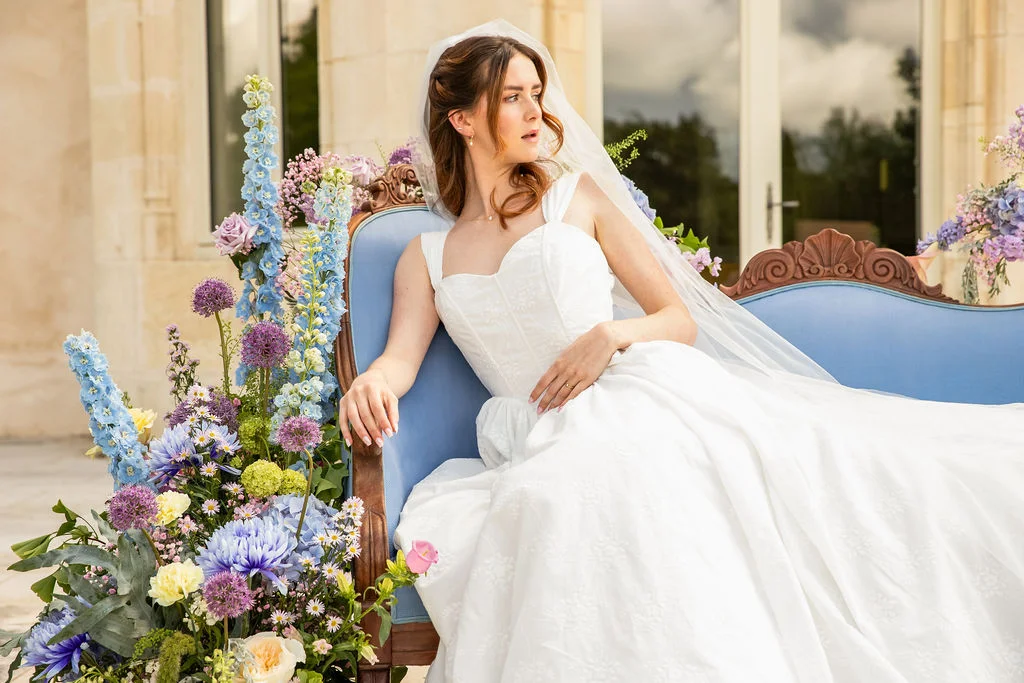 Une mariée en robe blanche et voile posant sur un canapé bleu entouré de fleurs pastel dans un cadre romantique.