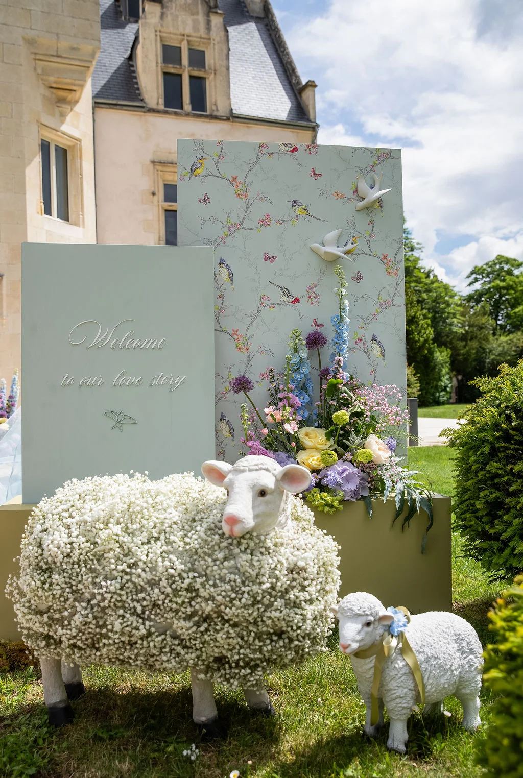 Décoration romantique avec moutons en fleurs et panneau de bienvenue devant un château sous un ciel bleu.
