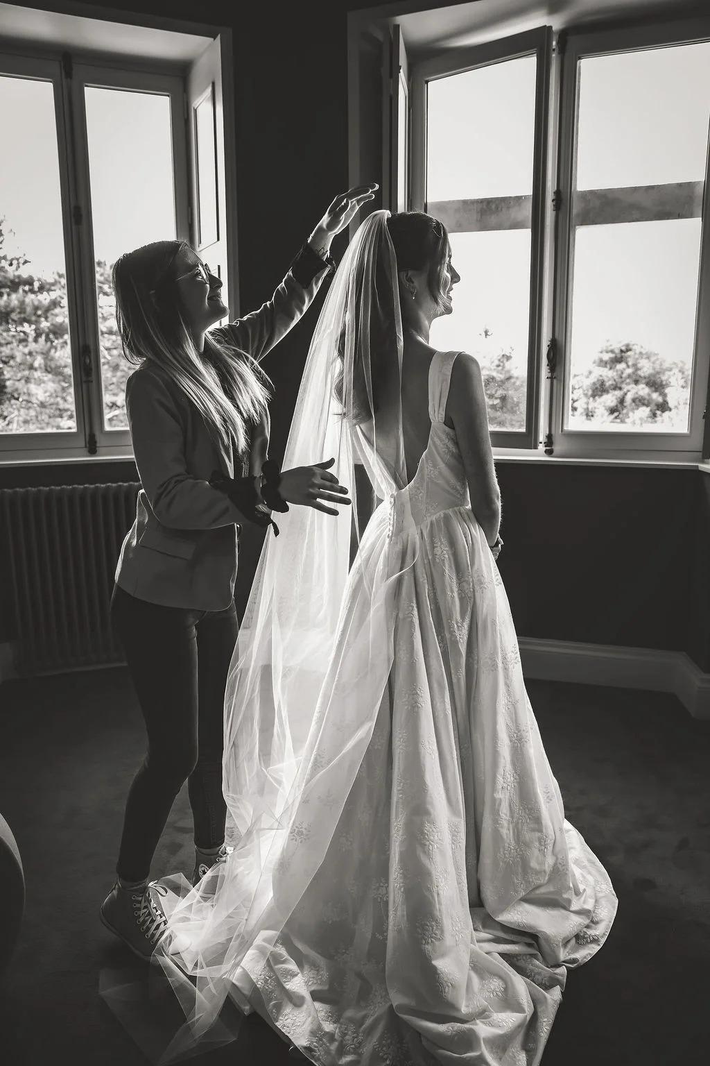 Photo noir et blanc d'une mariée dont le voile est ajusté par une assistante près d'une fenêtre lumineuse.