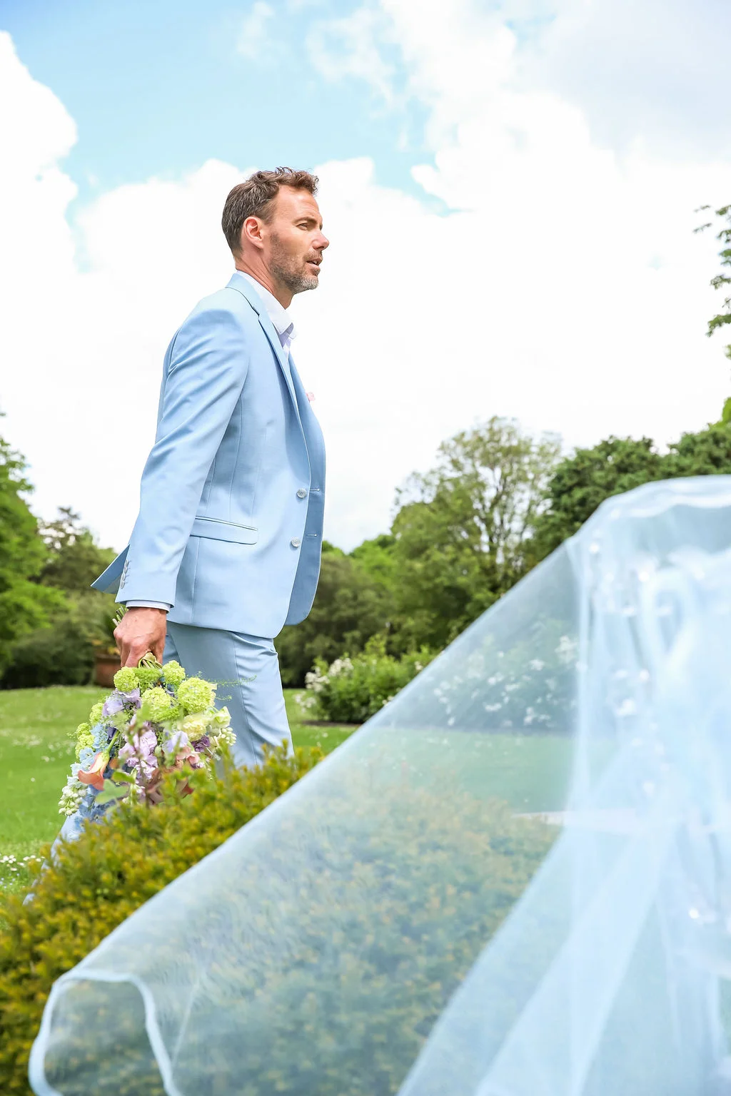 Un homme en costume bleu ciel tenant un bouquet coloré dans un jardin verdoyant sous un ciel lumineux.