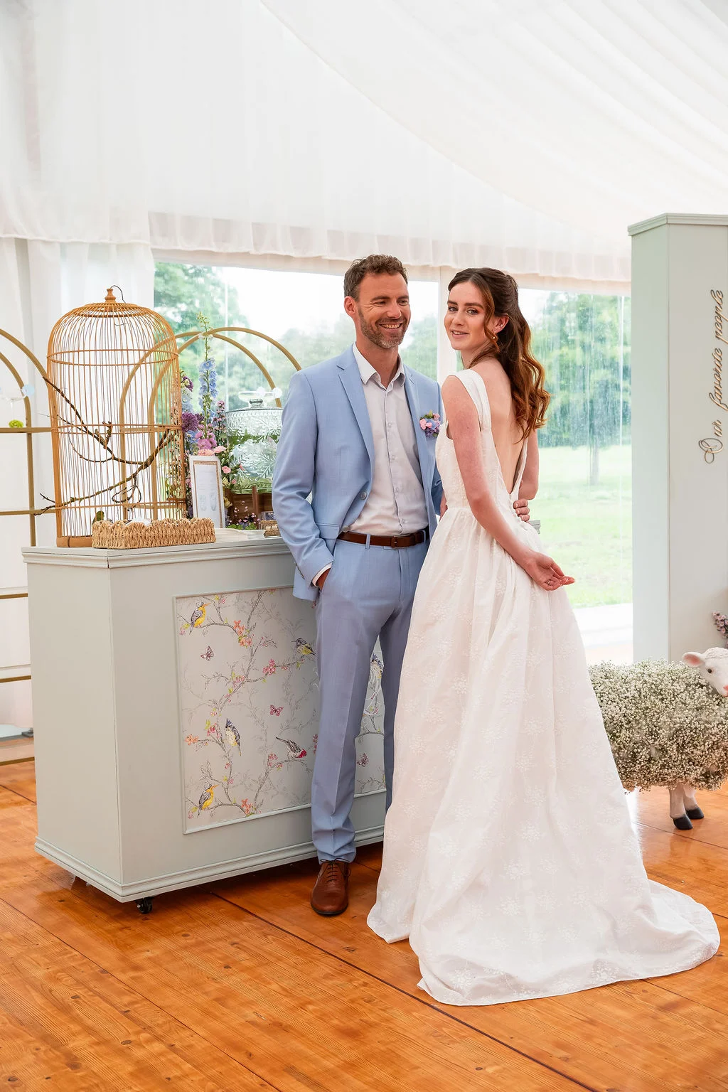 Couple marié souriant dans un décor romantique et lumineux avec un costume bleu ciel et une robe blanche fluide.