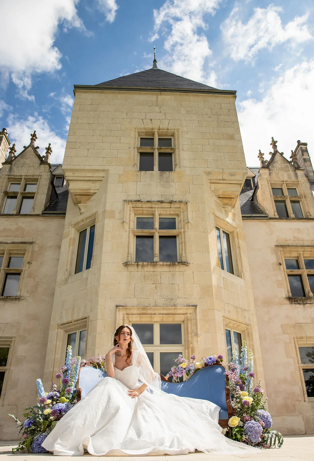 Mariée en robe blanche et voile assise sur un canapé bleu fleuri devant un château en pierre sous un ciel bleu.