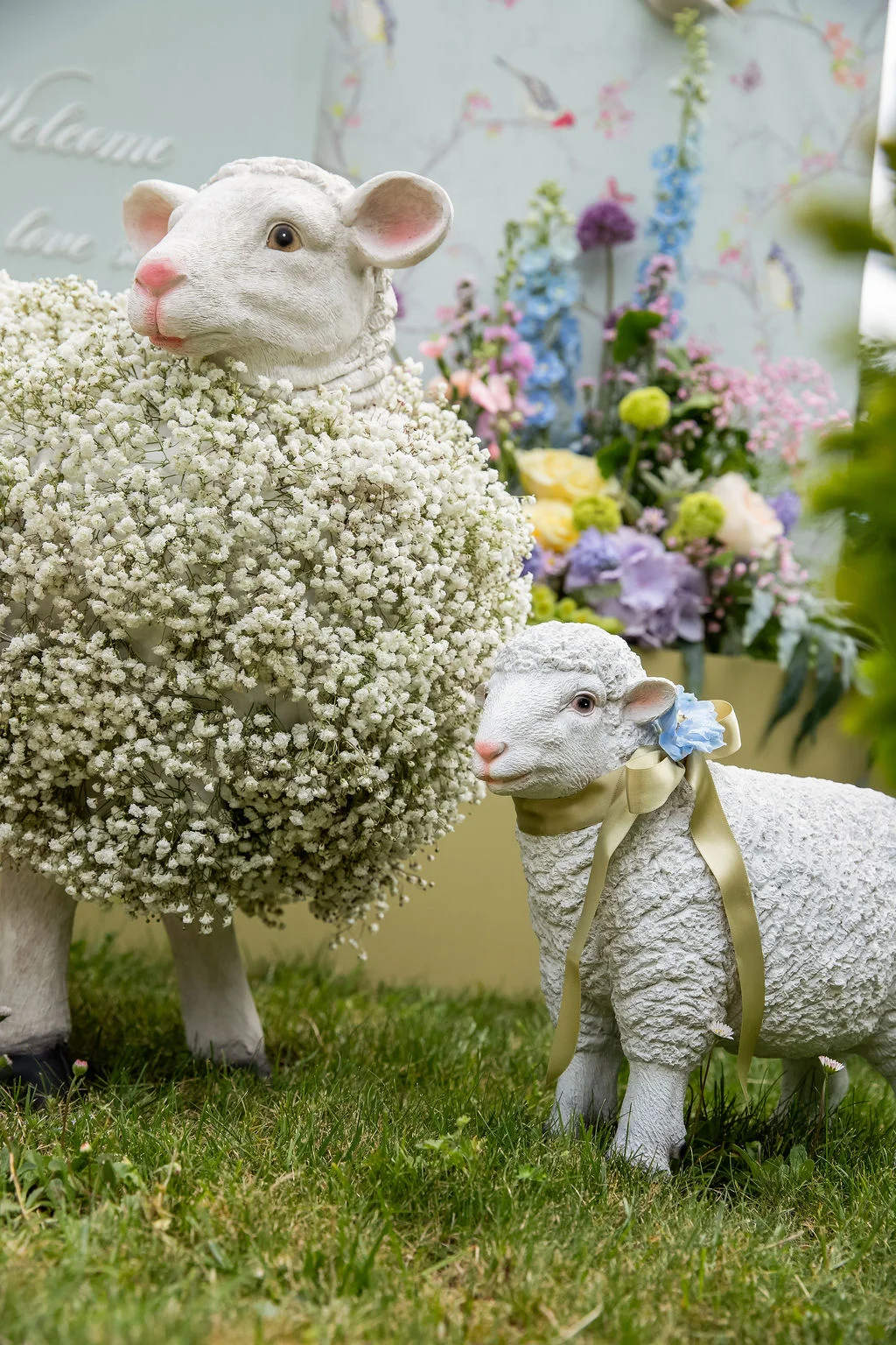 Deux figurines de moutons blancs, l'une ornée de gypsophile, devant un arrangement floral pastel et romantique.
