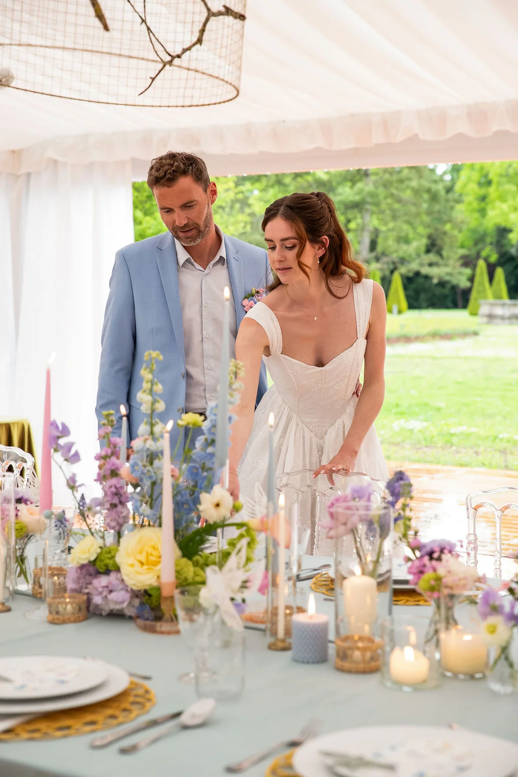Un couple de mariés ajuste la décoration florale pastel d'une table romantique sous un chapiteau blanc.
