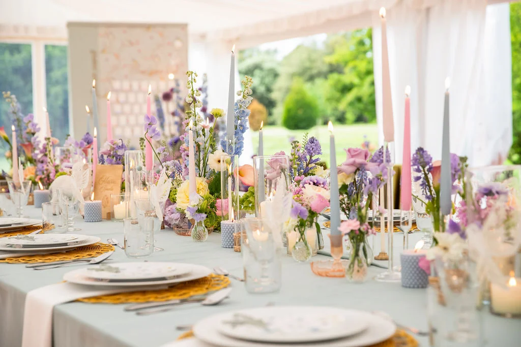 Table de mariage romantique aux tons pastel avec compositions florales violettes et bougies effilées sous un chapiteau.