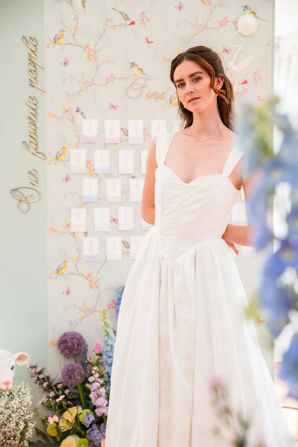Mariée en robe blanche romantique devant un mur floral pastel et des compositions de fleurs printanières.
