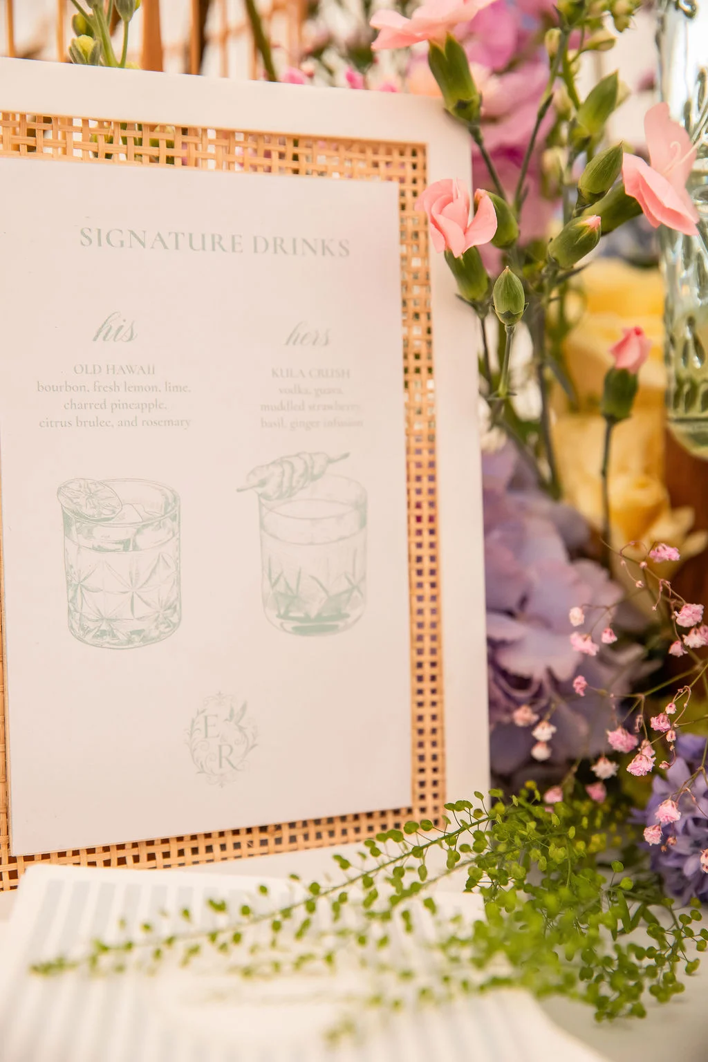 Menu de boissons signature dans un cadre en rotin, entouré de fleurs pastel pour un mariage romantique.