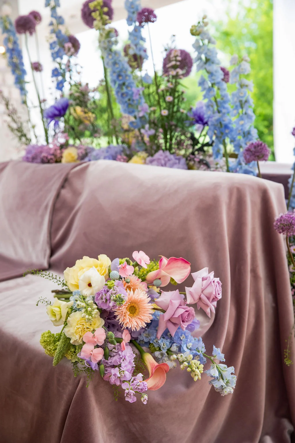 Bouquet de fleurs pastel posé sur un canapé en velours mauve, devant une composition florale bleue et violette.