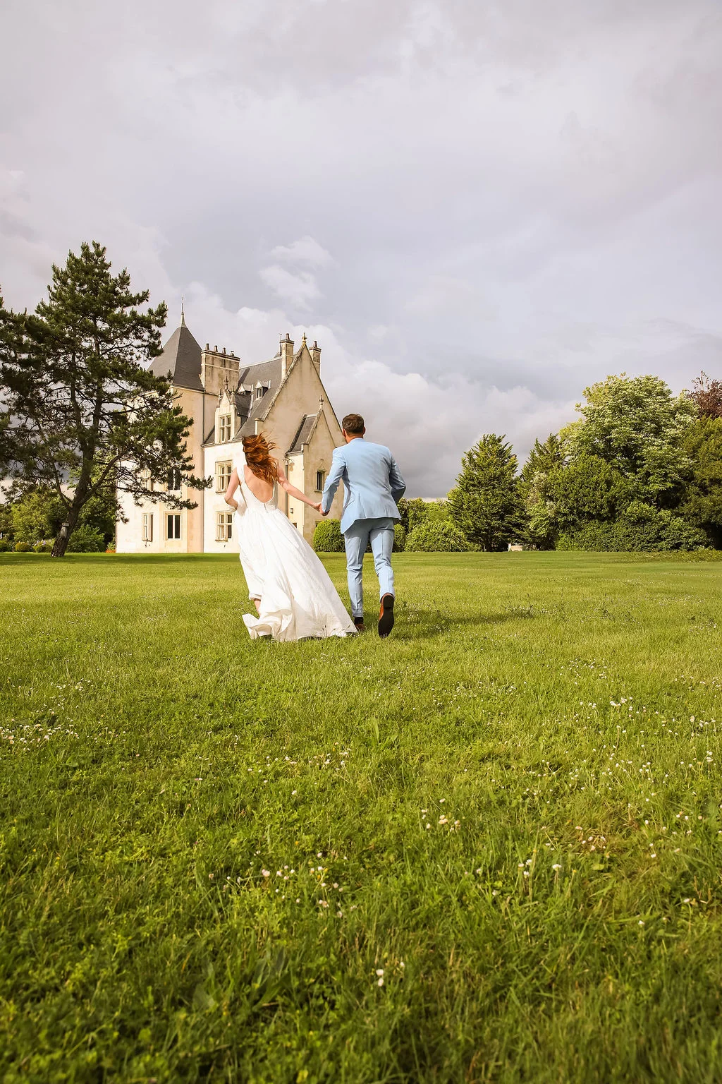 Jeunes mariés courant main dans la main vers un château blanc dans un vaste jardin verdoyant sous un ciel nuageux.