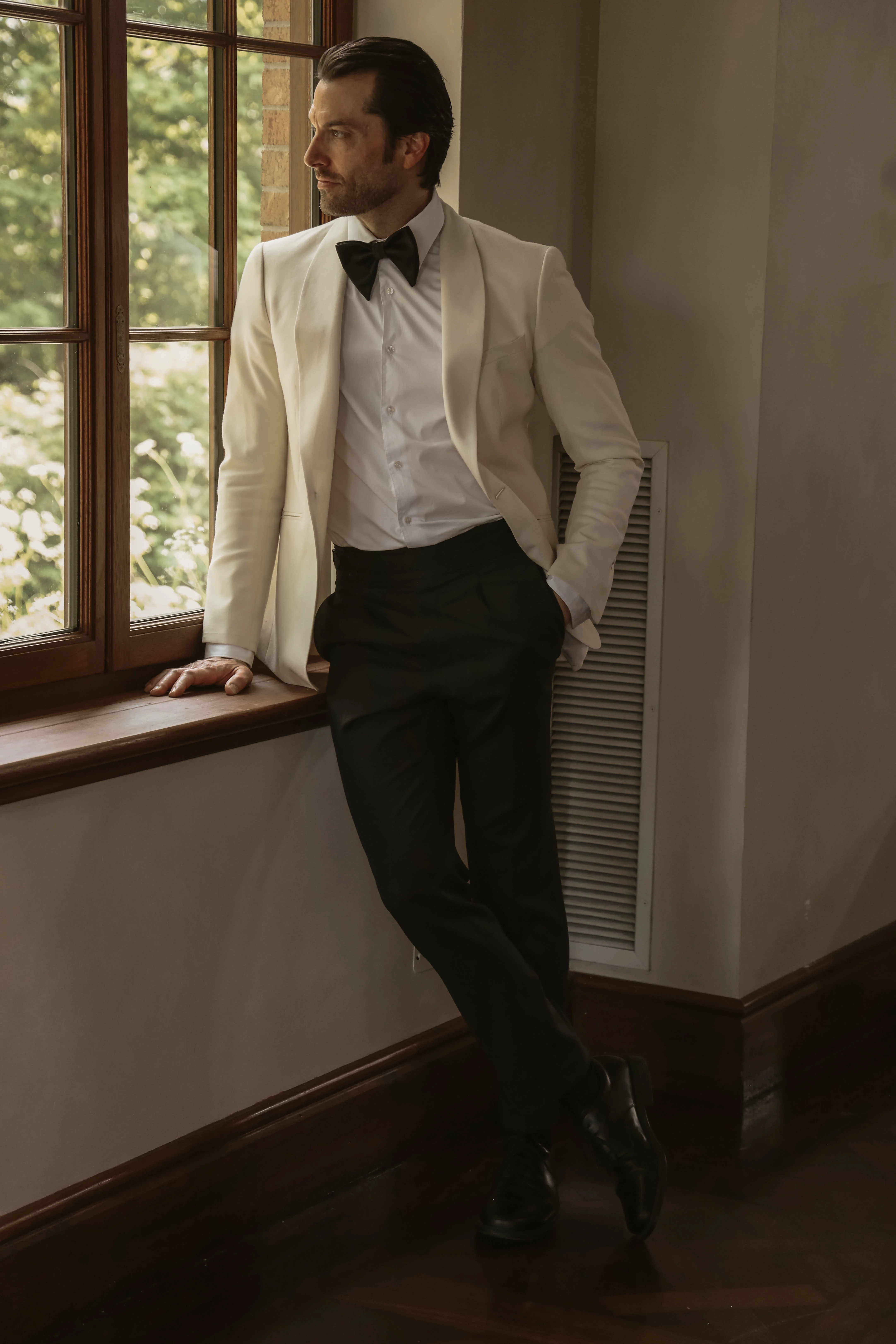 Homme élégant en smoking blanc et pantalon noir, posant près d'une fenêtre dans une ambiance lumineuse et minimaliste.