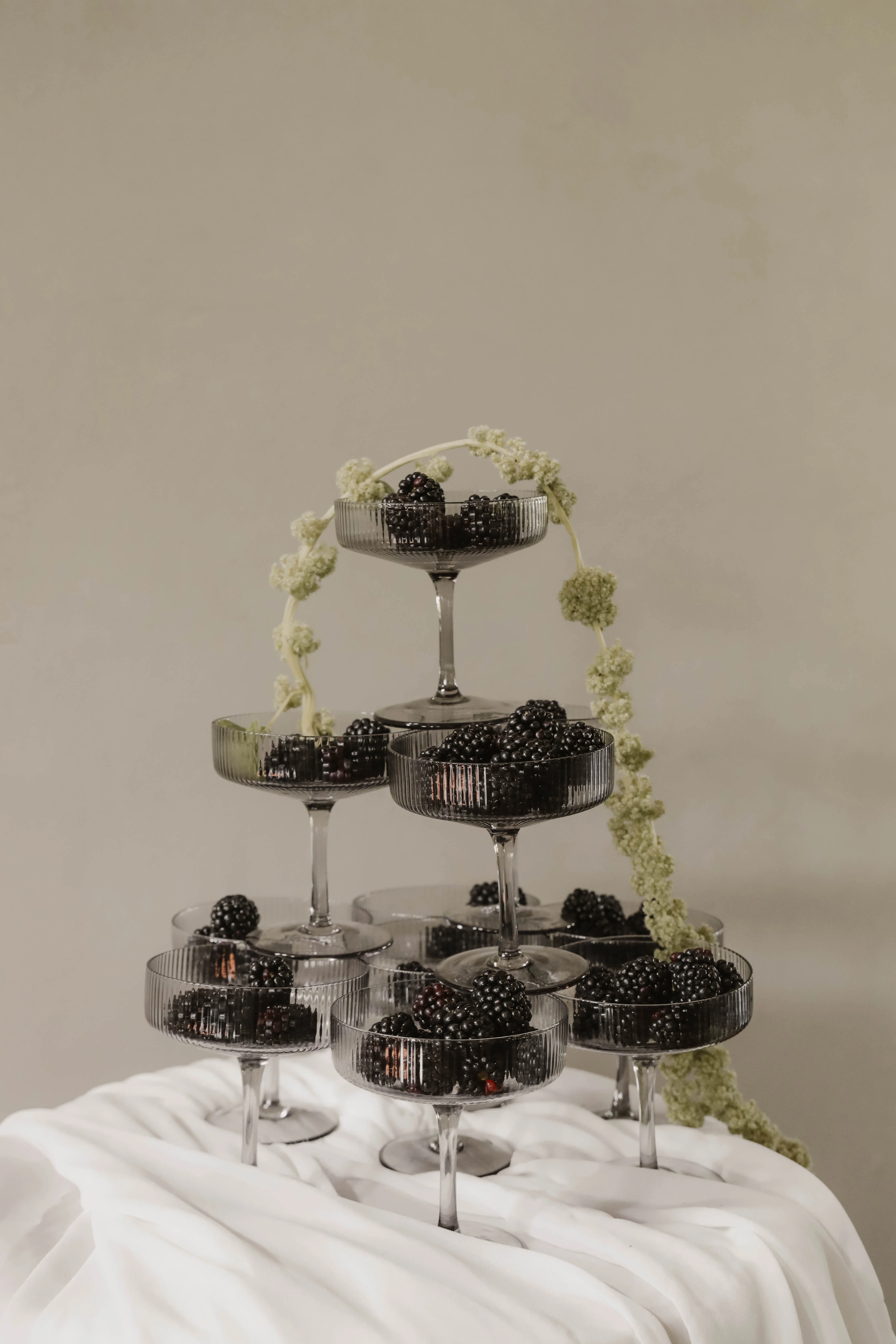 Pyramide de coupes en verre fumé remplies de mûres, décorée d'une branche fleurie sur un drapé blanc minimaliste.