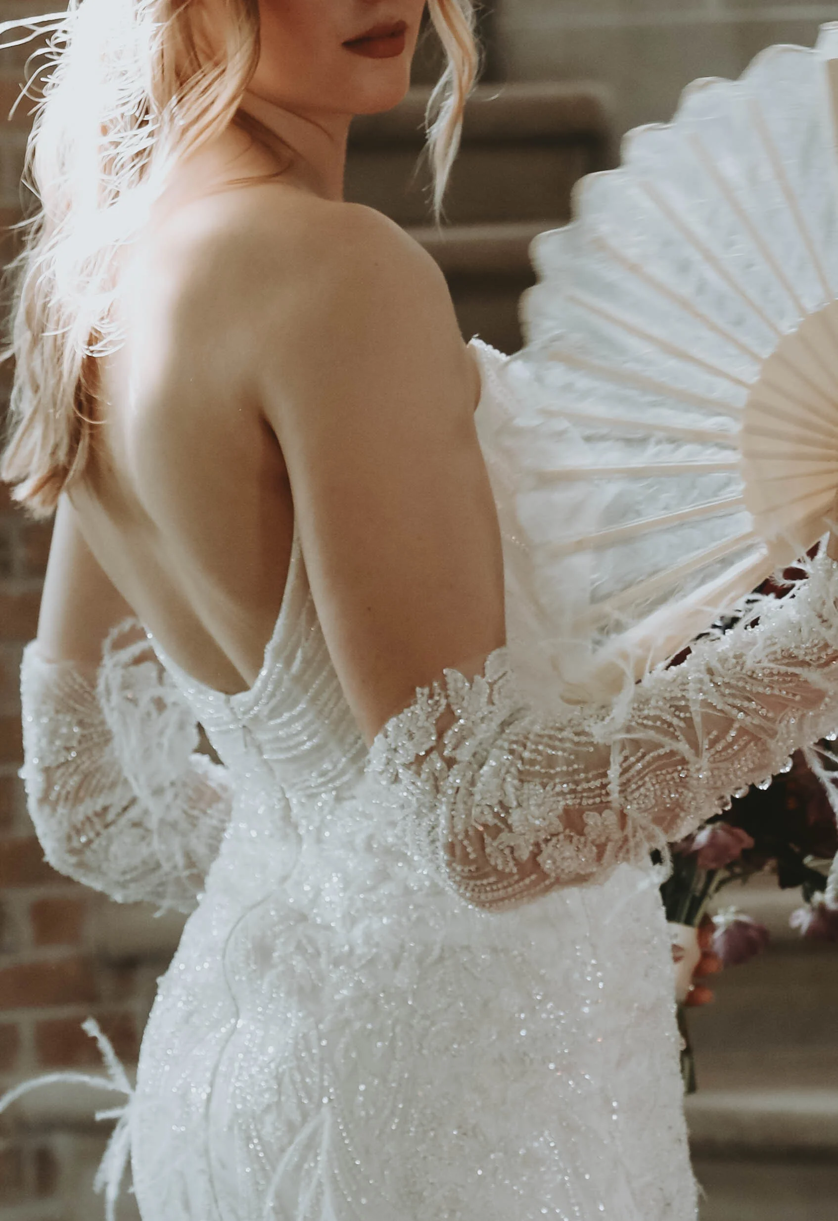 Mariée de dos en robe blanche scintillante et gants en dentelle, tenant un éventail blanc dans une ambiance romantique.