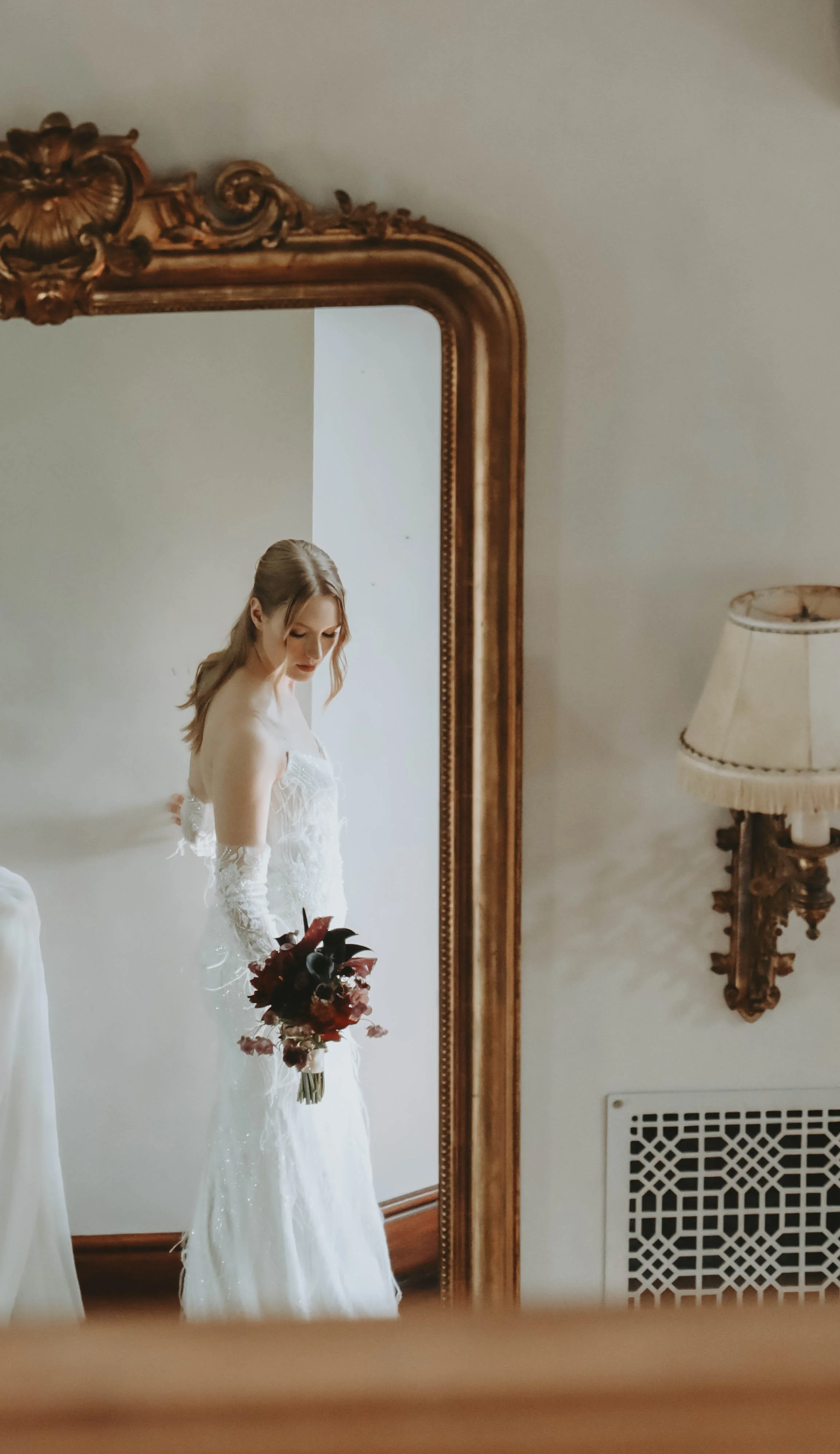 Une mariée en robe blanche et gants longs se reflète dans un miroir doré avec un bouquet rouge sombre.