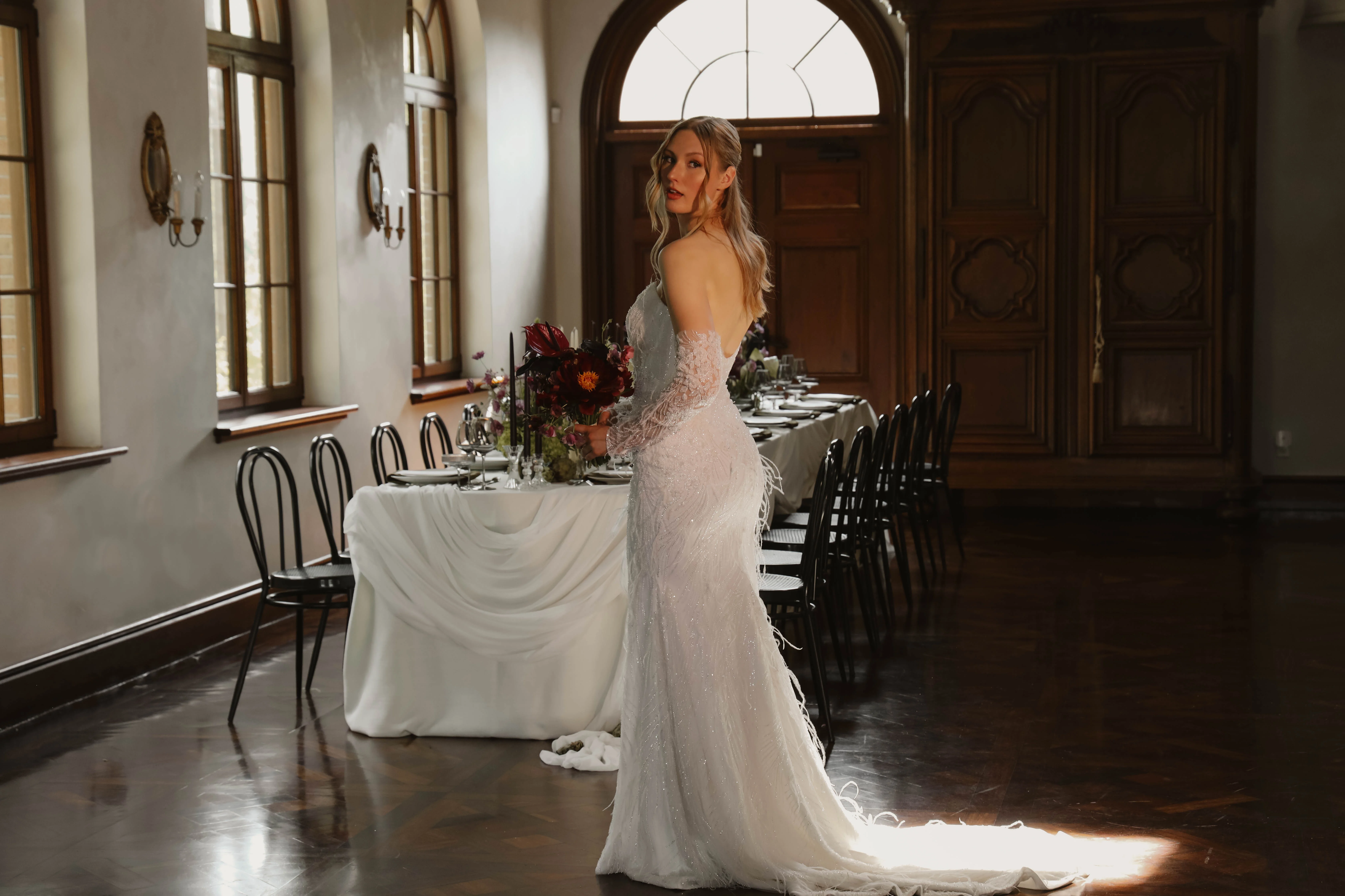 Mariée en robe sirène blanche scintillante posant dans une salle élégante avec une table minimaliste et fleurie.