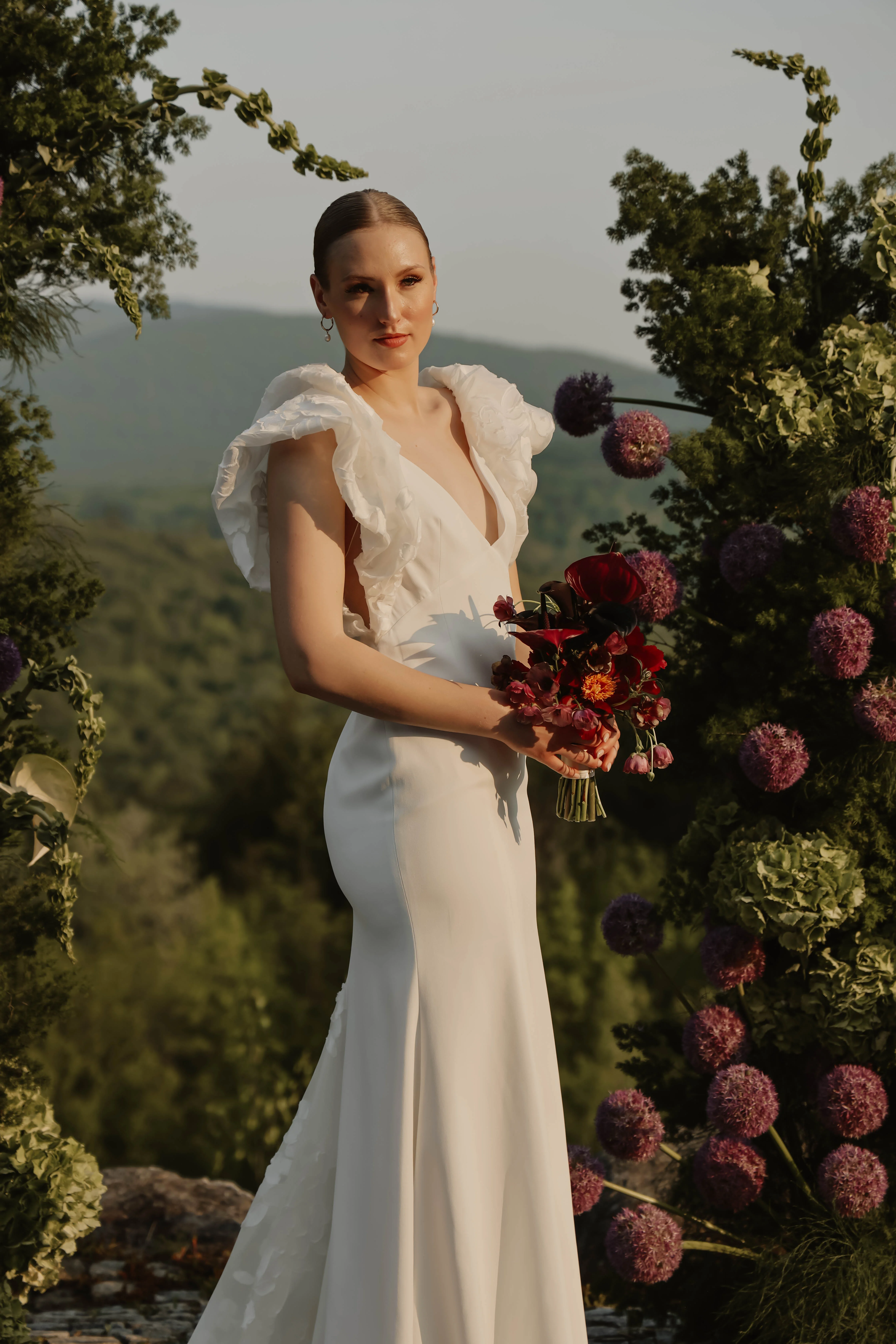 Mariée en robe blanche minimaliste avec volants, tenant un bouquet rouge sombre devant une arche florale naturelle.