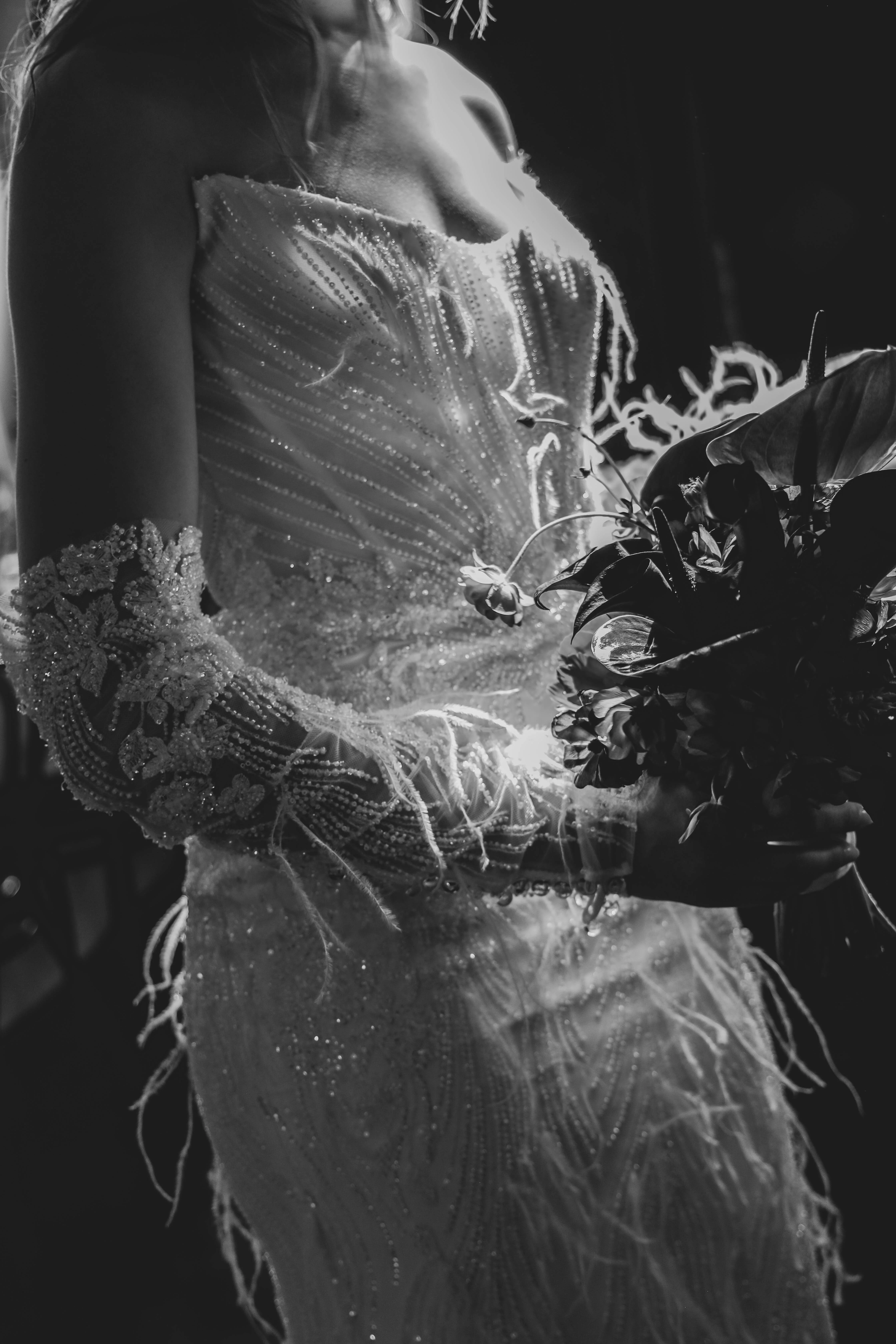 Portrait en noir et blanc d'une mariée en robe scintillante avec gants assortis, tenant un bouquet floral sombre.