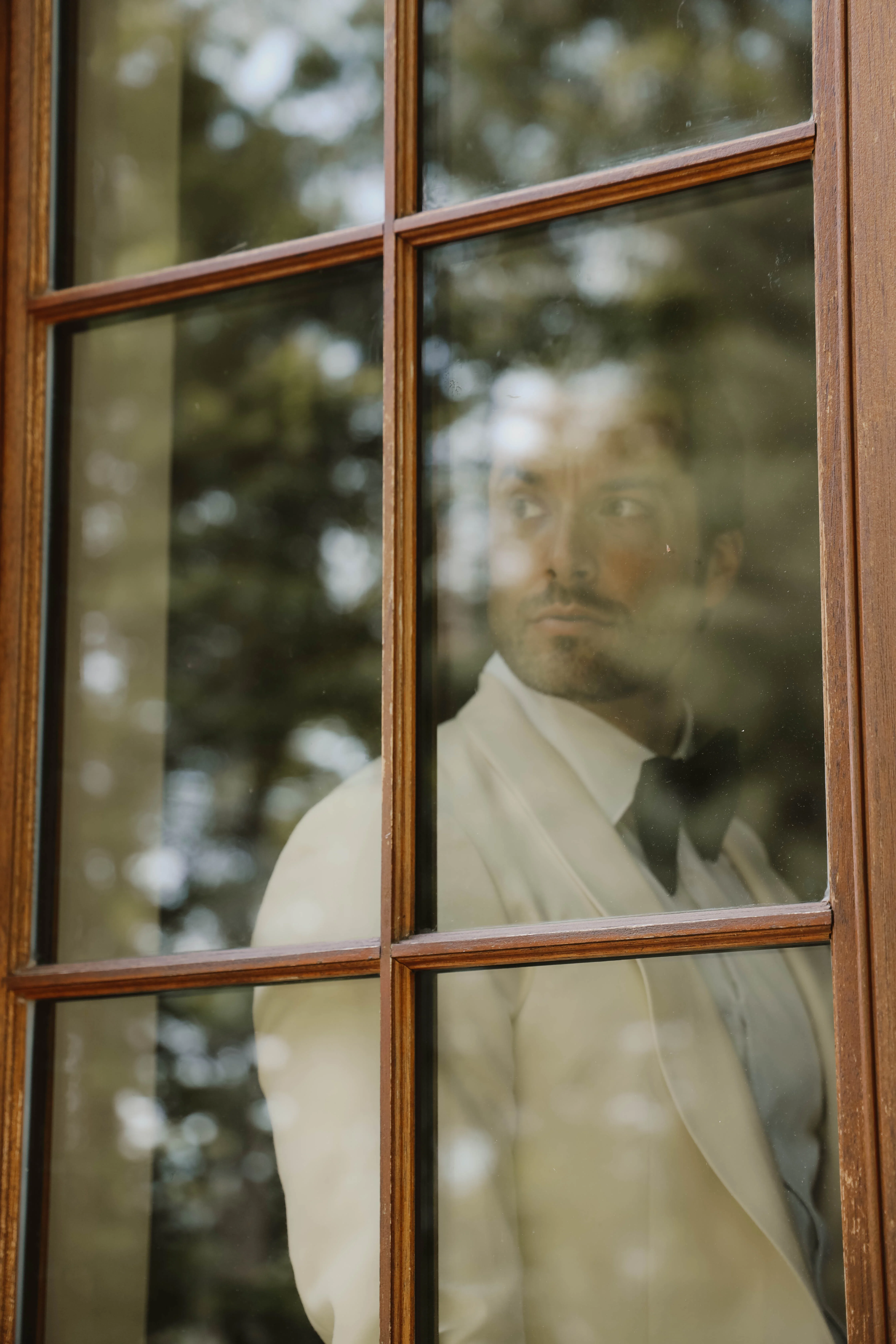 Reflet d'un marié en smoking blanc à travers une fenêtre en bois, ambiance romantique et minimaliste.