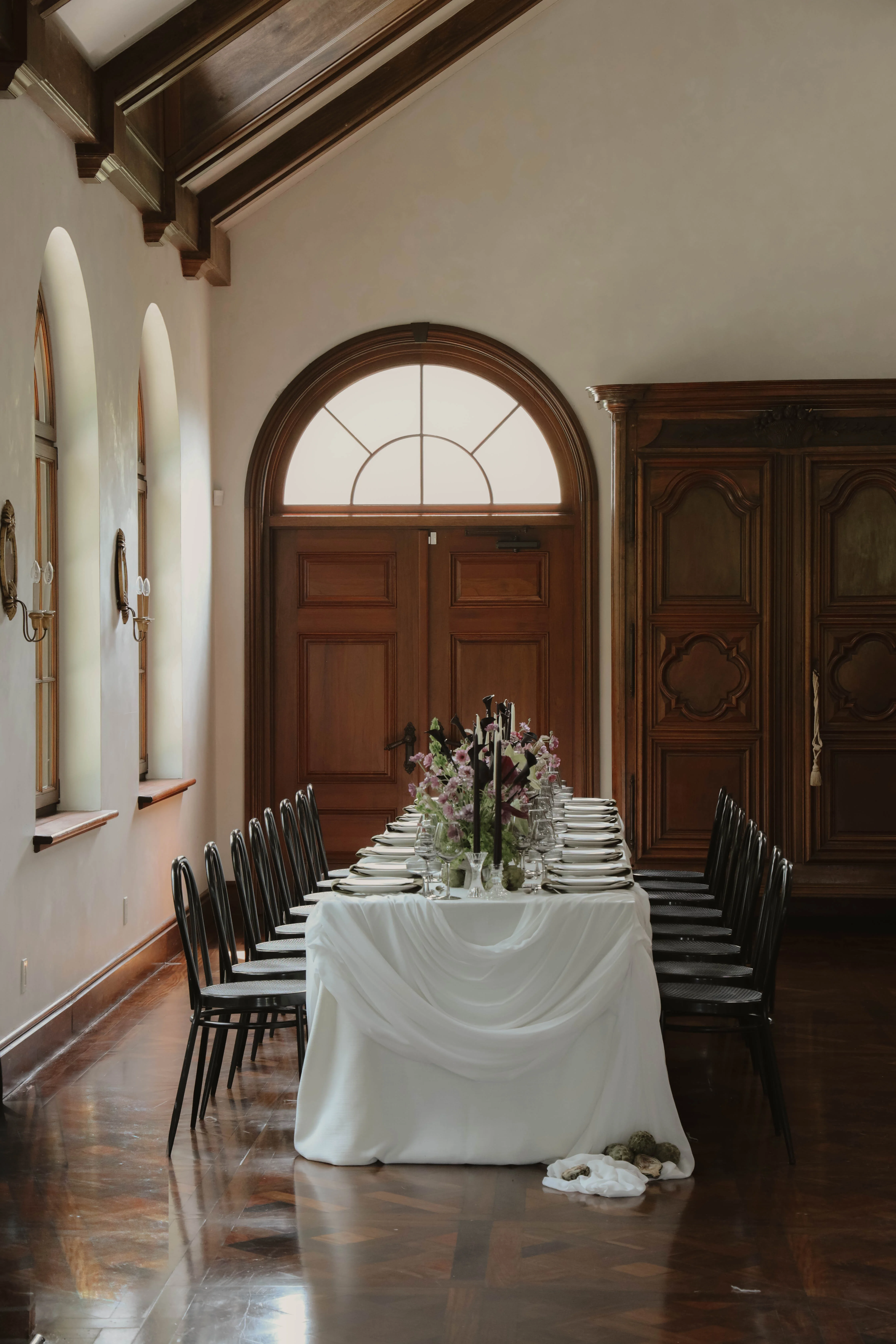 Table de banquet minimaliste nappée de blanc avec fleurs et chaises noires dans une salle aux boiseries chaleureuses.