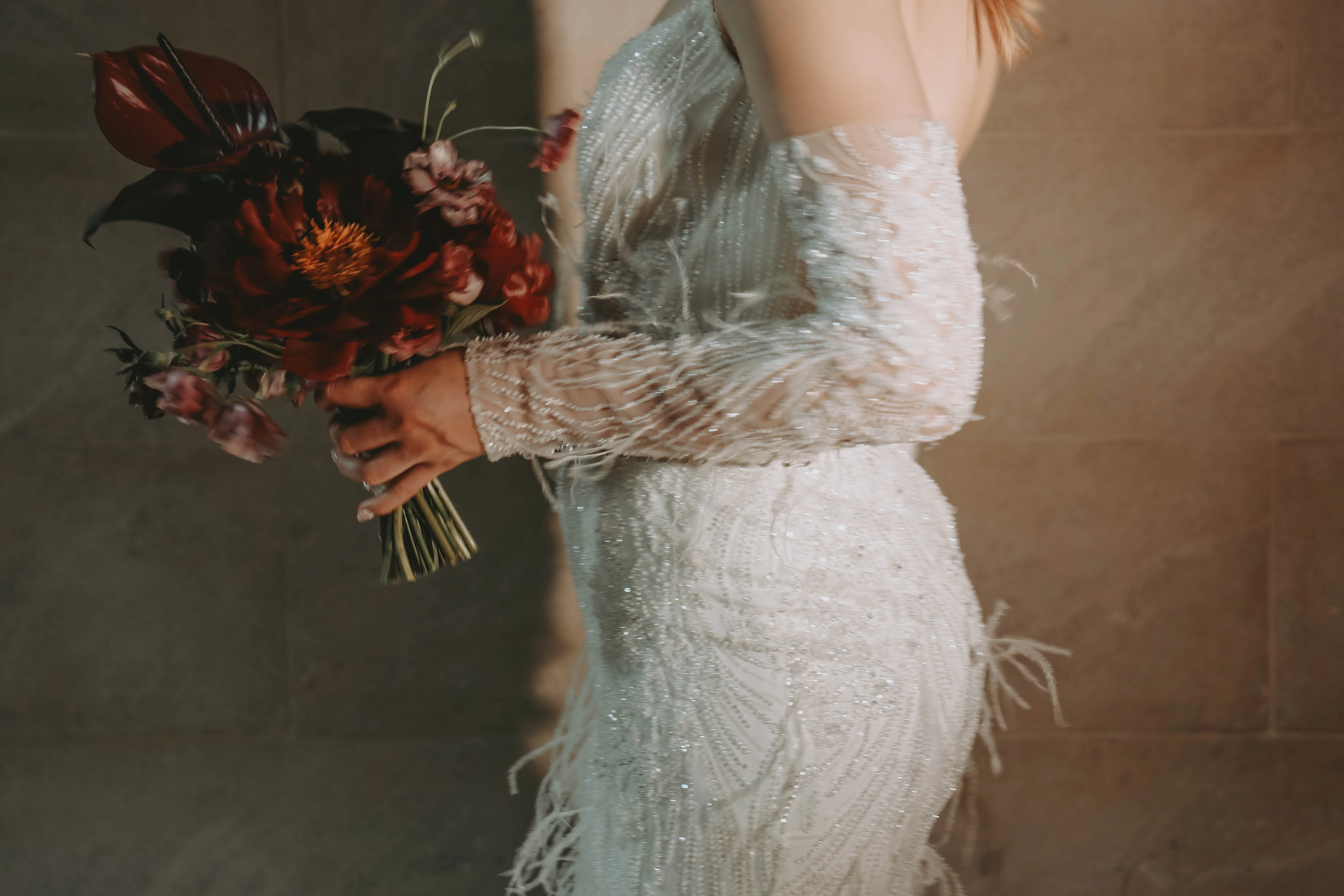 Mariée en robe blanche scintillante tenant un bouquet de fleurs rouge sombre sur fond gris minimaliste.