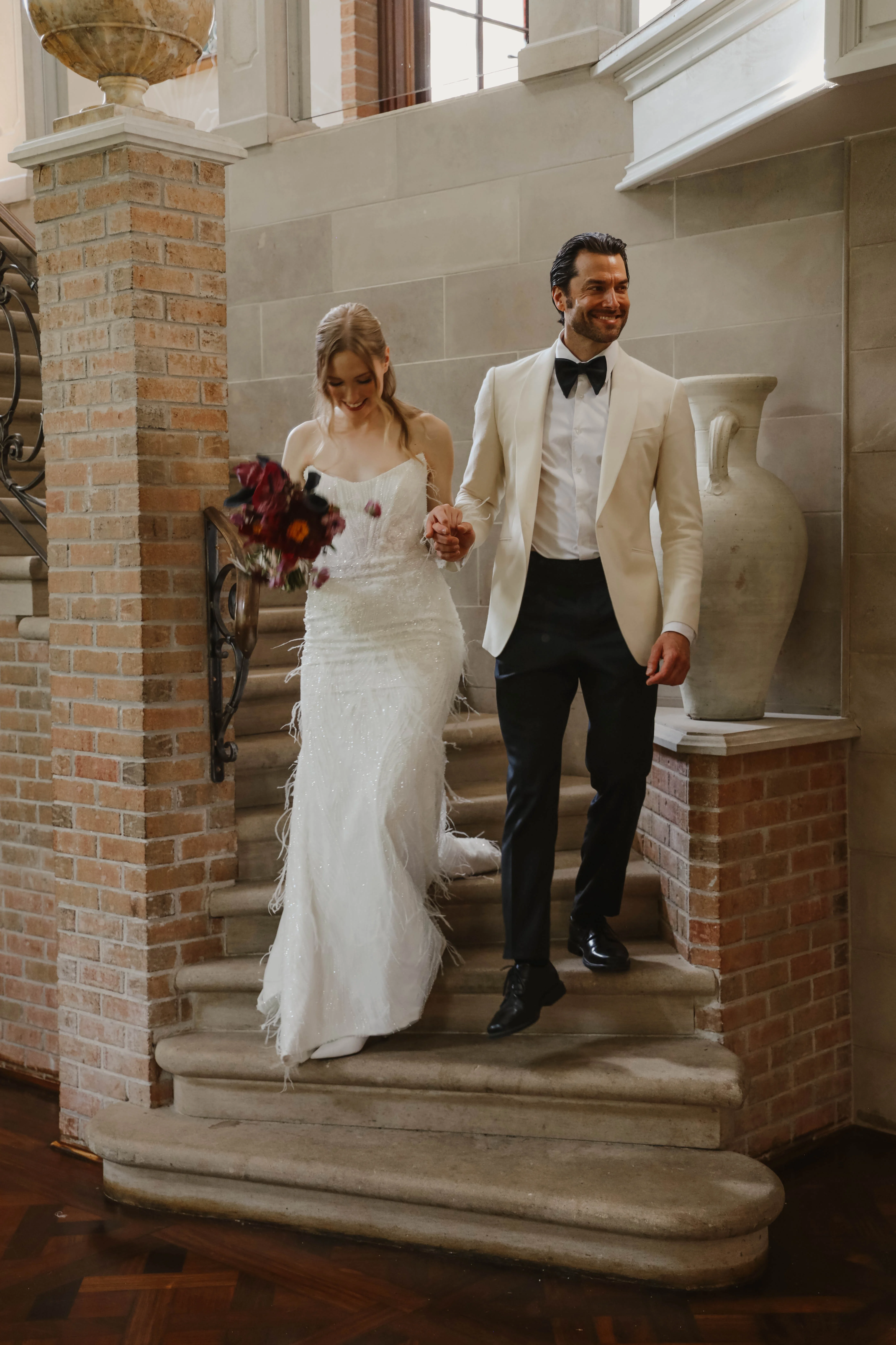 Jeunes mariés élégants descendant un escalier en pierre, elle en robe blanche scintillante et lui en smoking blanc et noir.