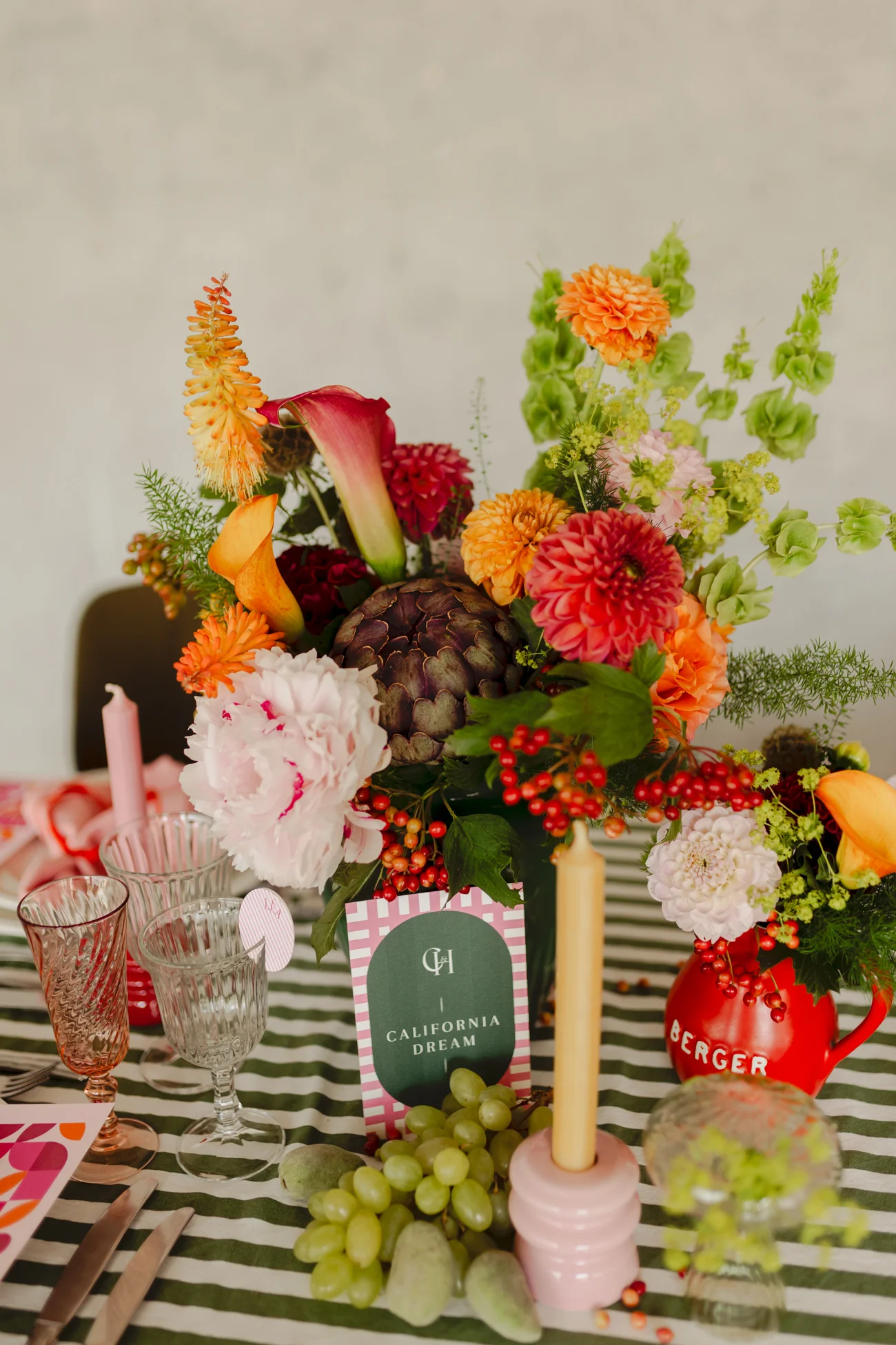 Composition florale colorée et rétro avec artichaut, raisins et bougies sur une nappe rayée verte et blanche.