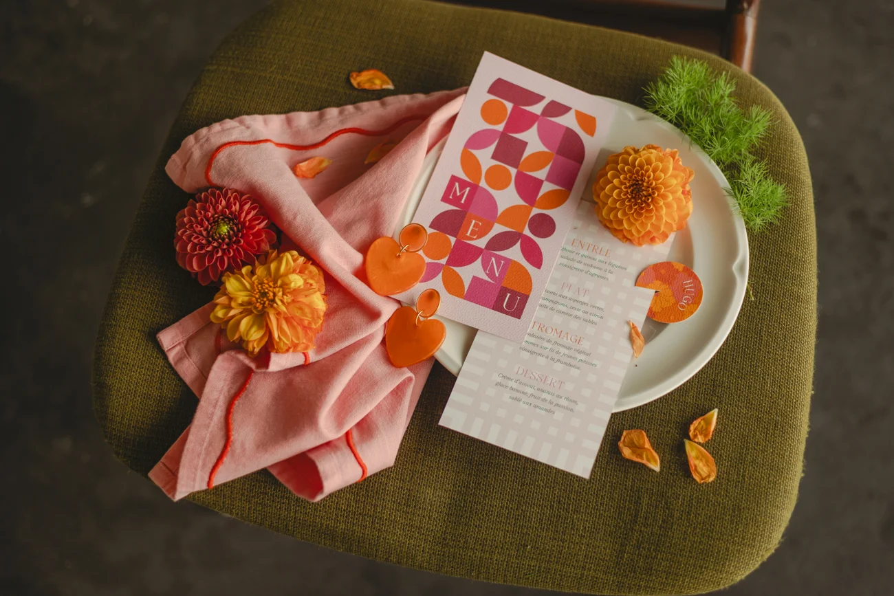 Mise en scène rétro avec menu géométrique, fleurs colorées et textiles roses sur un siège vert olive.