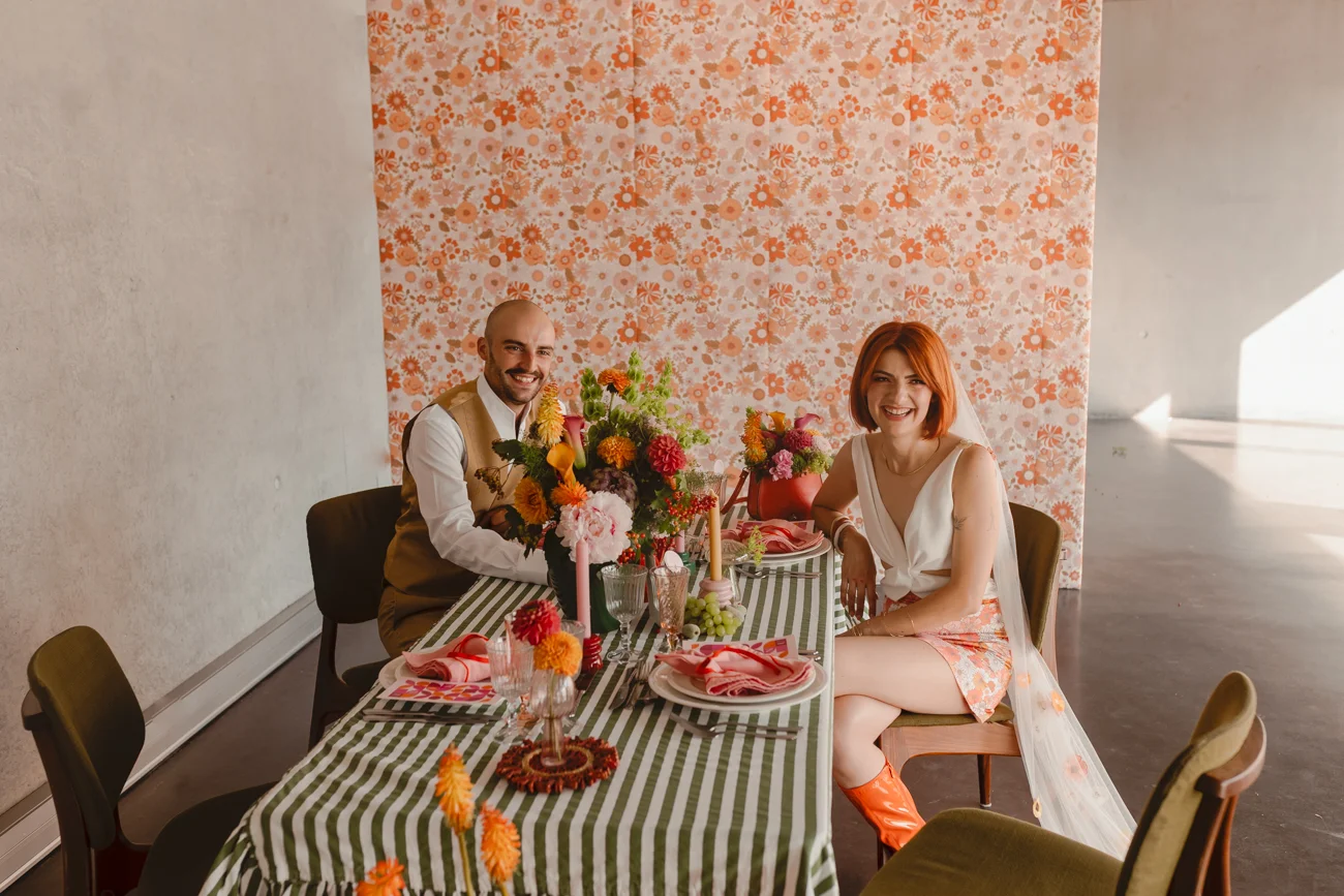 Couple souriant à une table festive rétro aux tons orange et vert, dans un décor vintage et coloré.