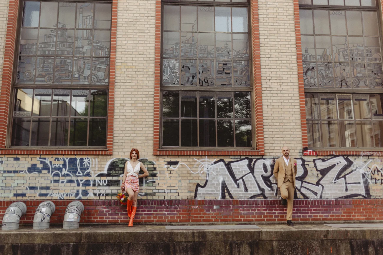 Couple au look rétro posant devant un mur de briques avec graffitis et de grandes fenêtres industrielles.