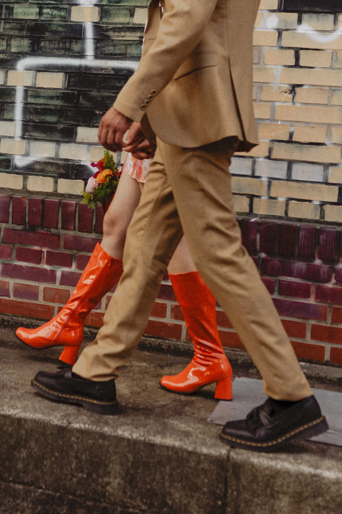 Couple en costume beige et bottes orange vernies marchant côte à côte devant un mur de briques.