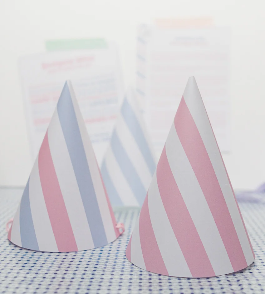 Trois chapeaux de fête coniques à rayures roses et bleues posés sur une nappe à carreaux bleus et blancs.