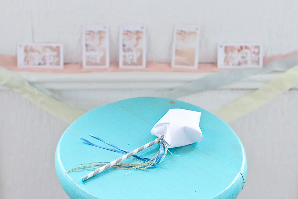 Baguette magique DIY avec pompon blanc et rubans bleus posée sur une table ronde turquoise.
