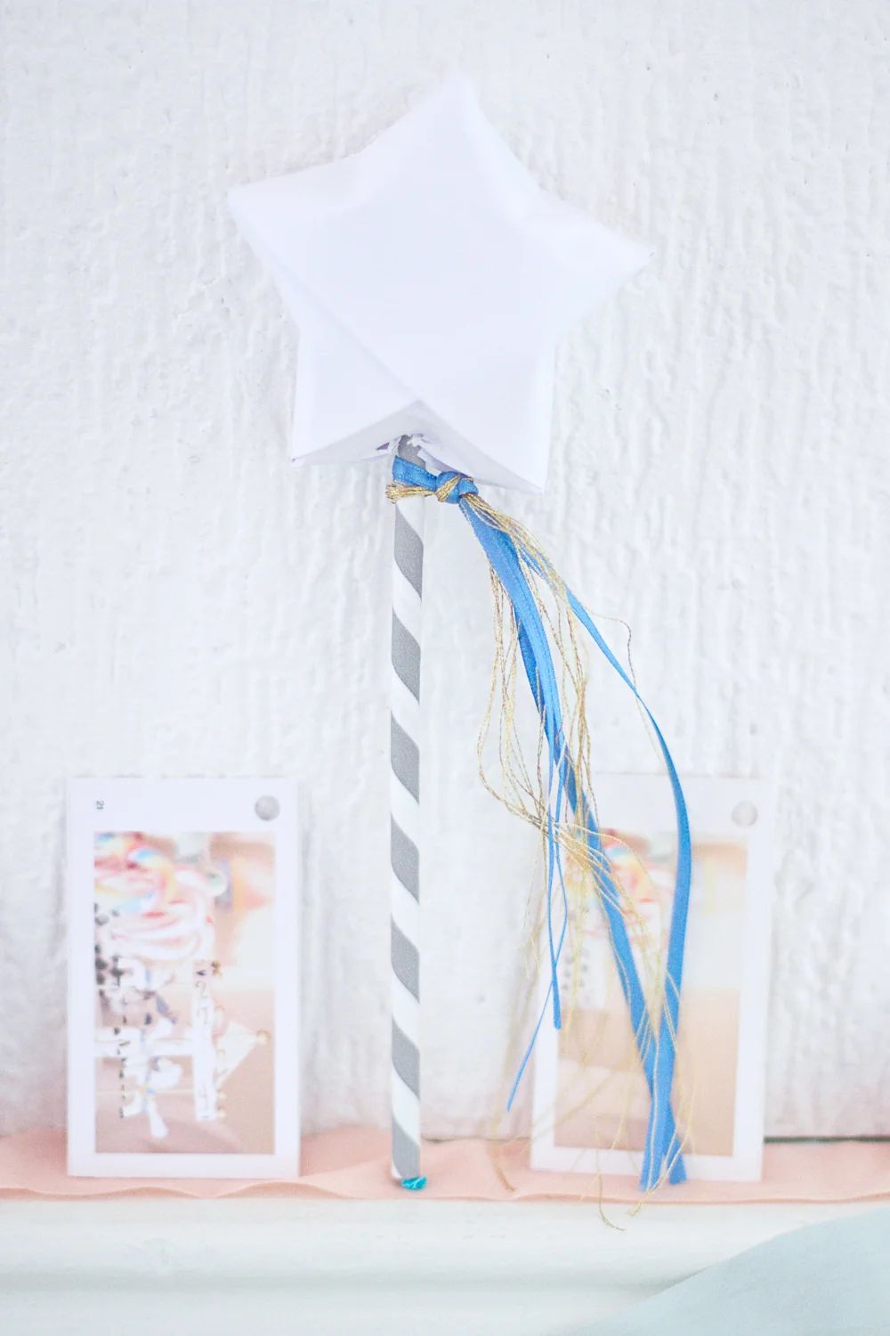 Baguette magique DIY avec une étoile blanche, un manche rayé gris et des rubans bleus et dorés sur fond blanc.